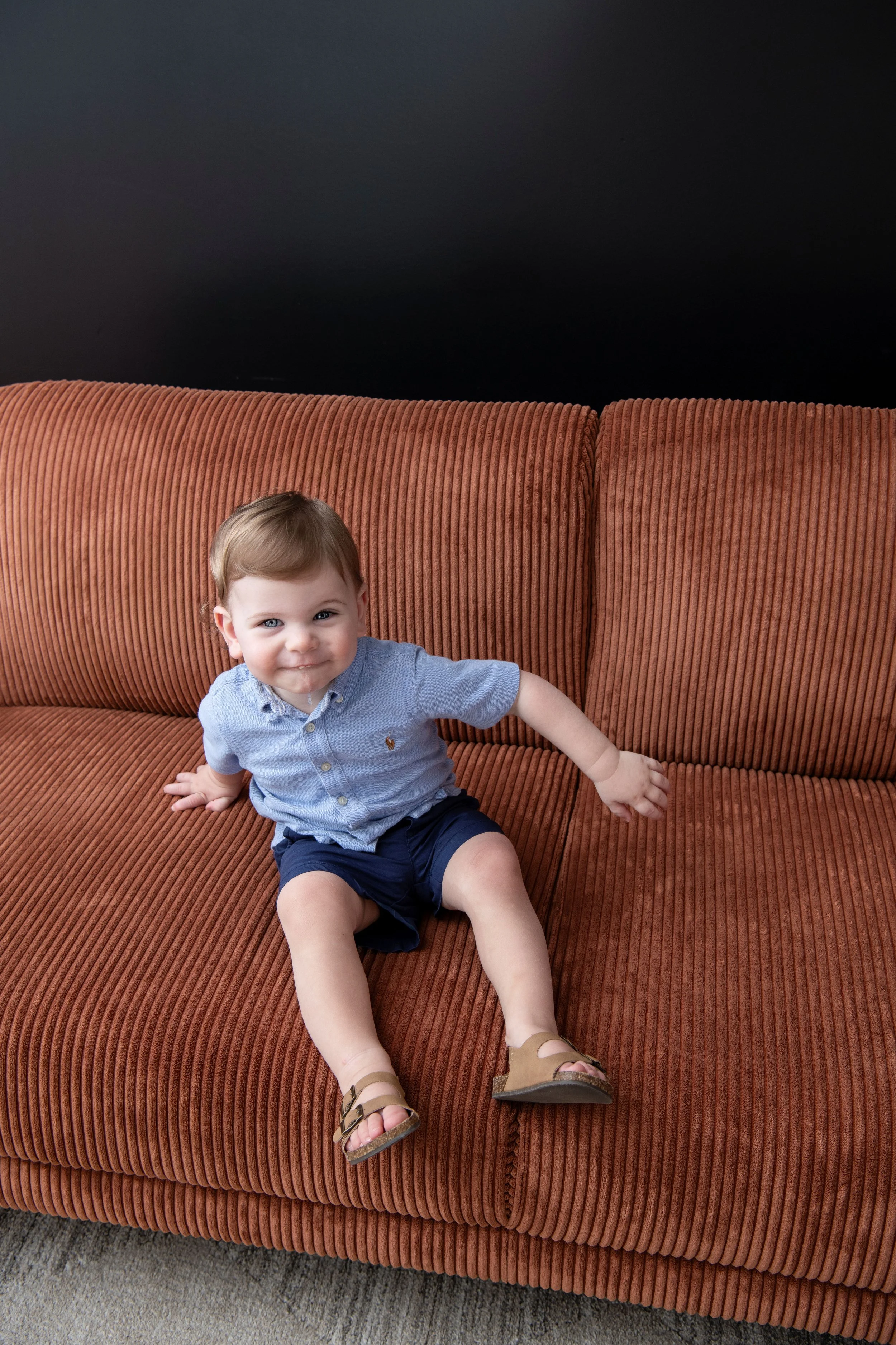 A young boy with brown hair, wearing a light blue button-up shirt with a small emblem, navy shorts, and tan sandals, sitting sideways on a rust-colored corduroy sofa with a dark background.
