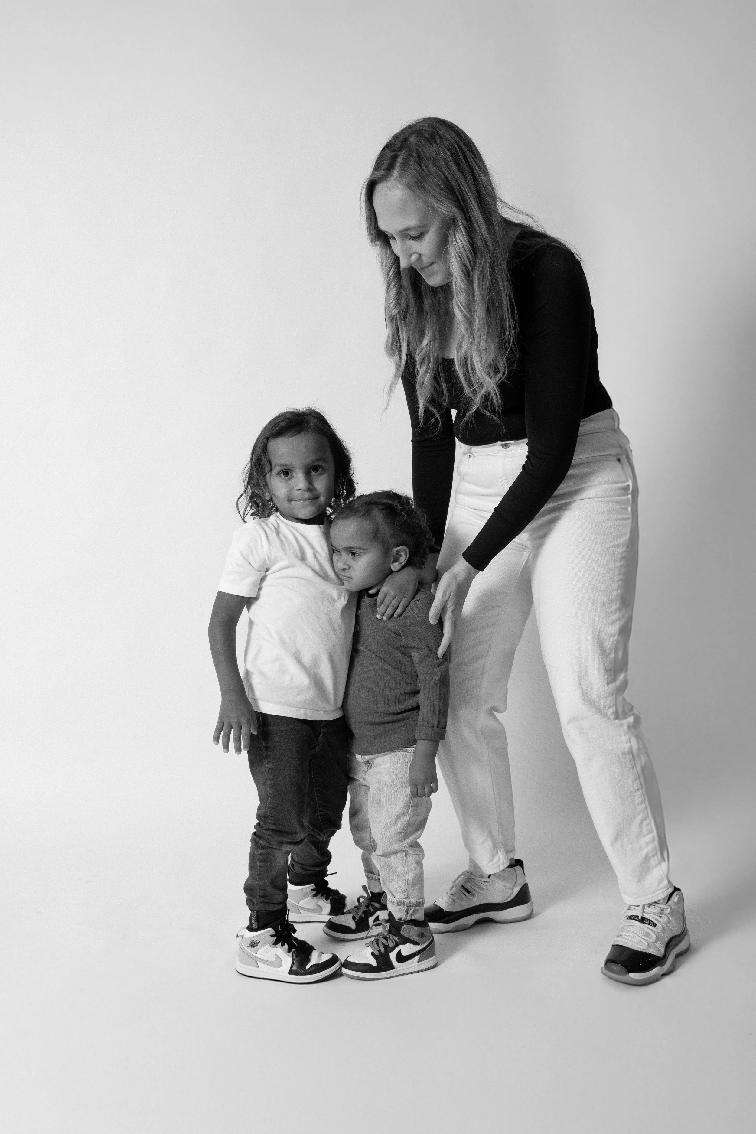 A woman with two young children standing close together against a plain background. The woman is bending down slightly, looking at the children, who are all wearing sneakers.