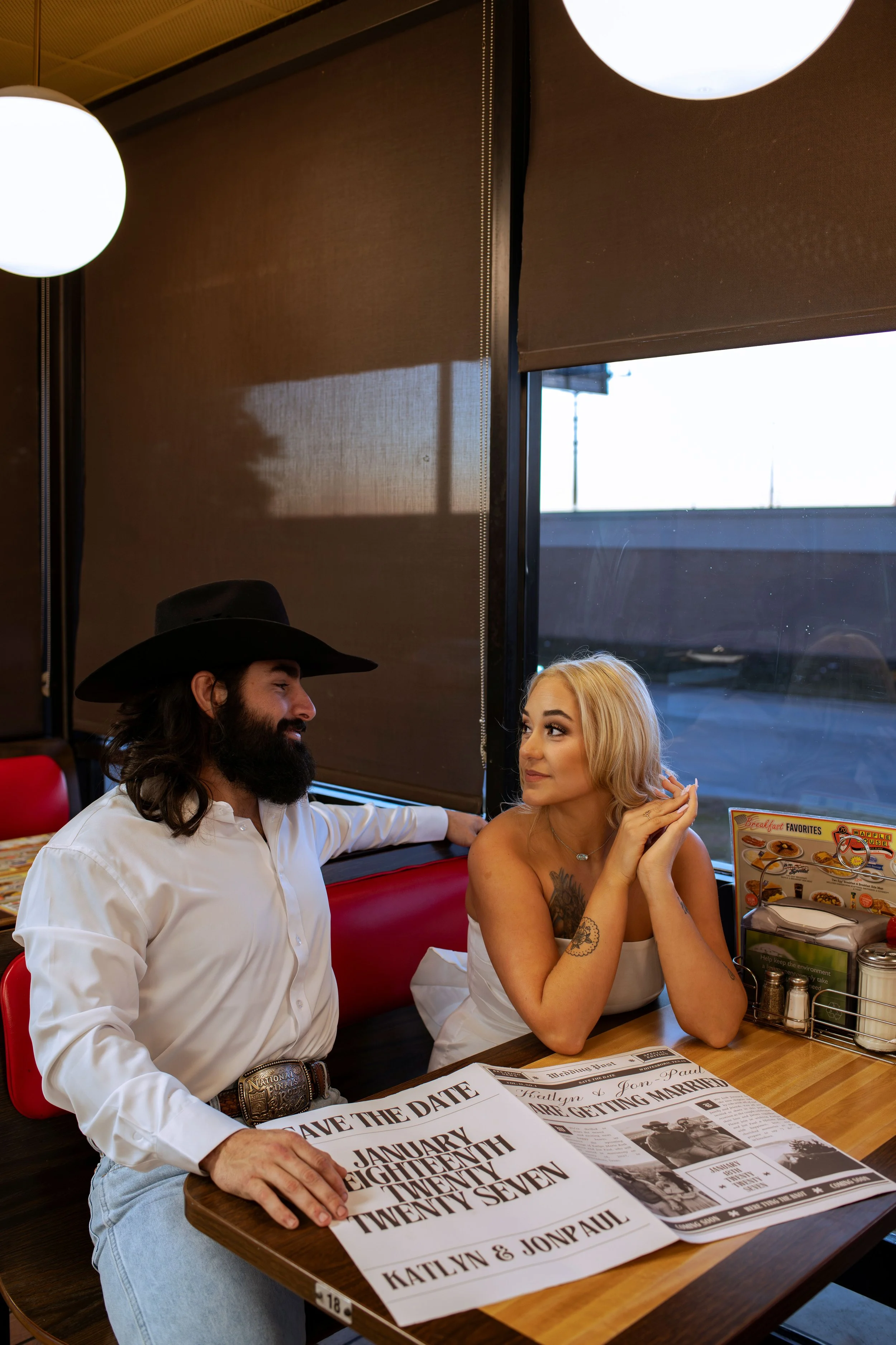 A man with a beard and long hair wearing a cowboy hat and a white shirt sits at a restaurant table, smiling at a woman with blonde hair, tattoos, and a white strapless top, who is looking at him with a smile. The table has papers, menus, and condimen
