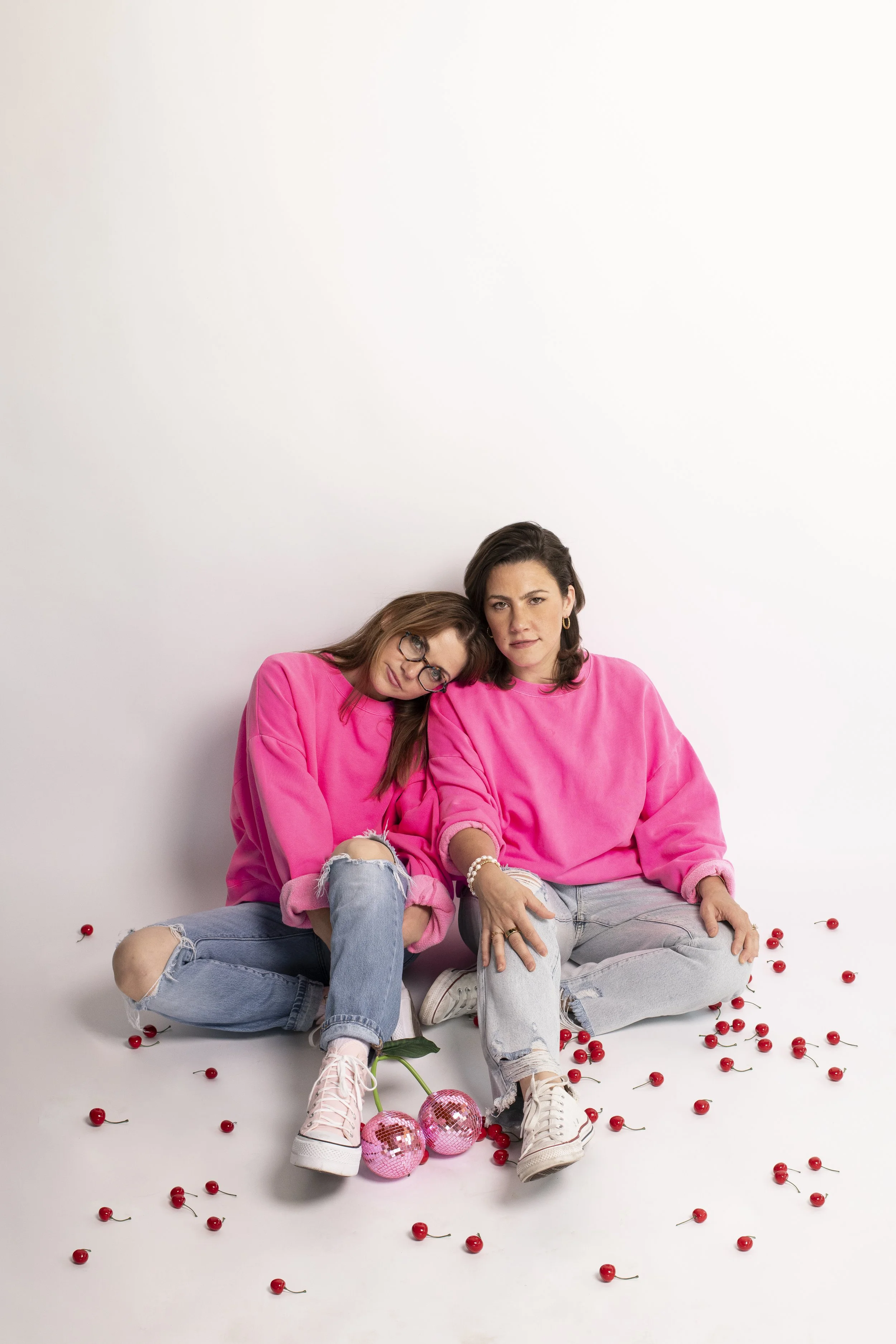 Two women sitting on the floor against a white background, wearing matching pink sweatshirts and ripped jeans, surrounded by scattered cherries and pink disco balls, with one wearing glasses and leaning her head on the other's shoulder.