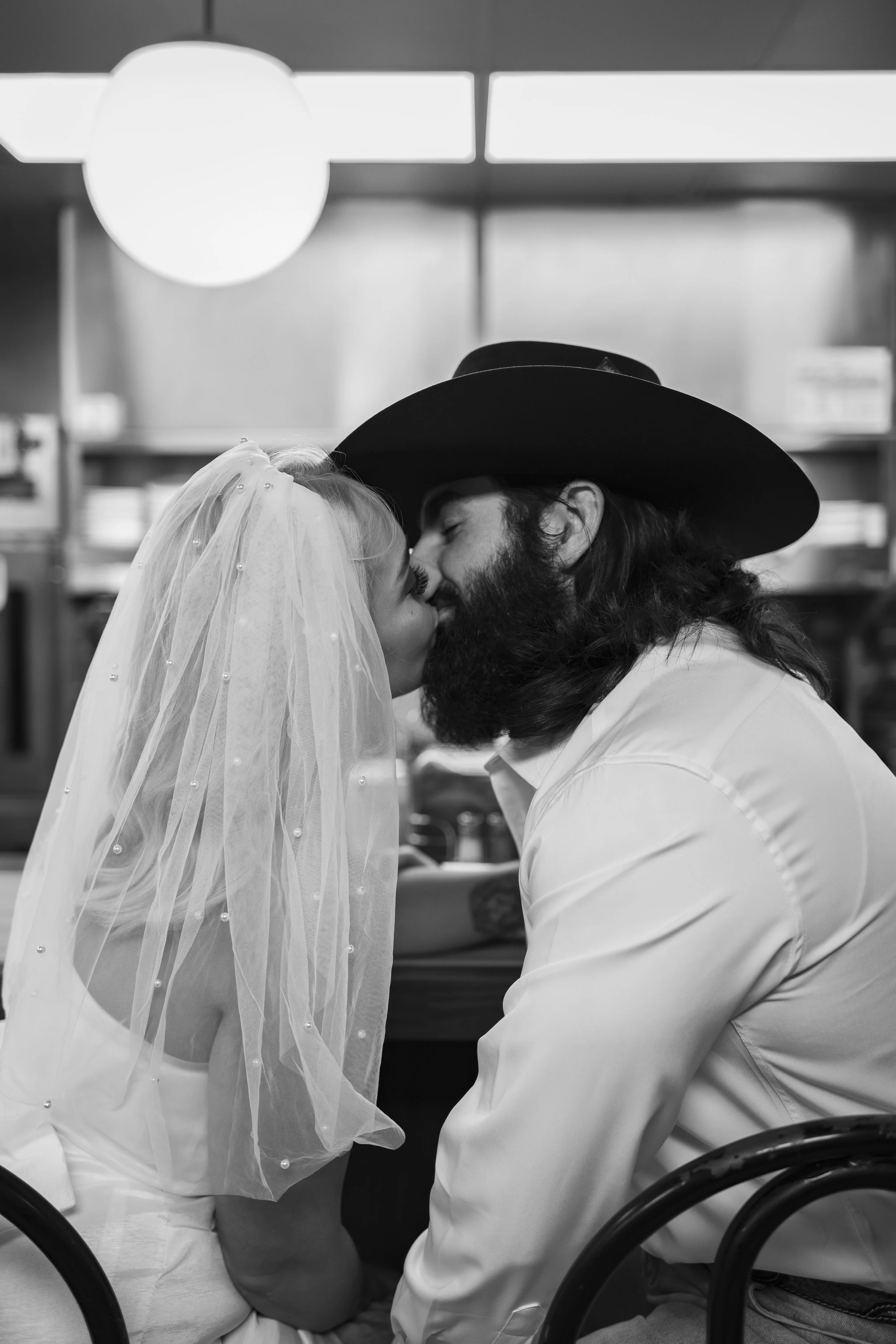 A black-and-white photo of a couple sharing a kiss. The woman is wearing a bridal veil with pearl embellishments, and the man is wearing a large cowboy hat and has a beard. They are seated at a table in an indoor setting, possibly a restaurant or café.