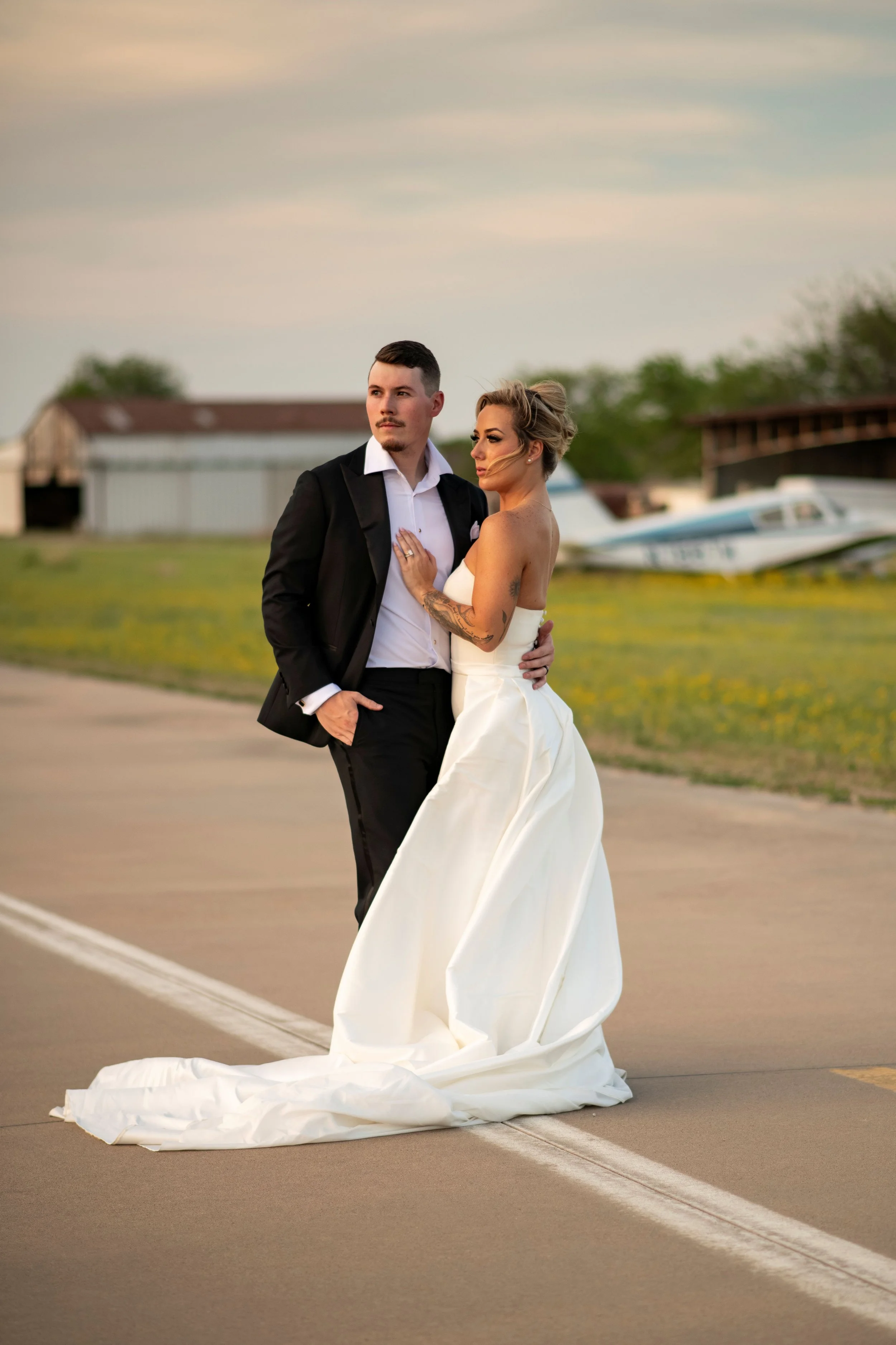 A newlywed couple dressed in wedding attire, standing on an airport tarmac near small planes and hangars, during sunset.