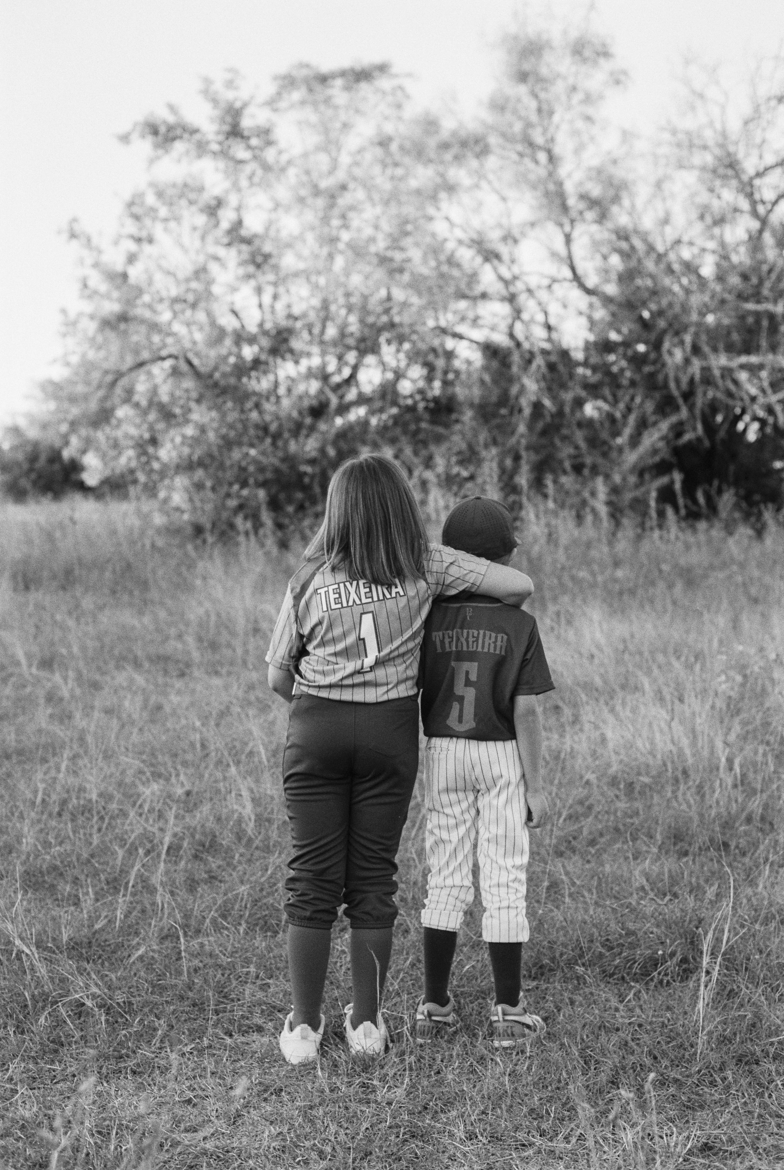 Two young children, a girl and a boy, walk together through a grassy field, with the girl resting her arm on the boy's shoulder. They wear baseball jerseys with the words 'TEXIERA' and numbers 1 and 5 on the back, matching striped pants, and sneakers