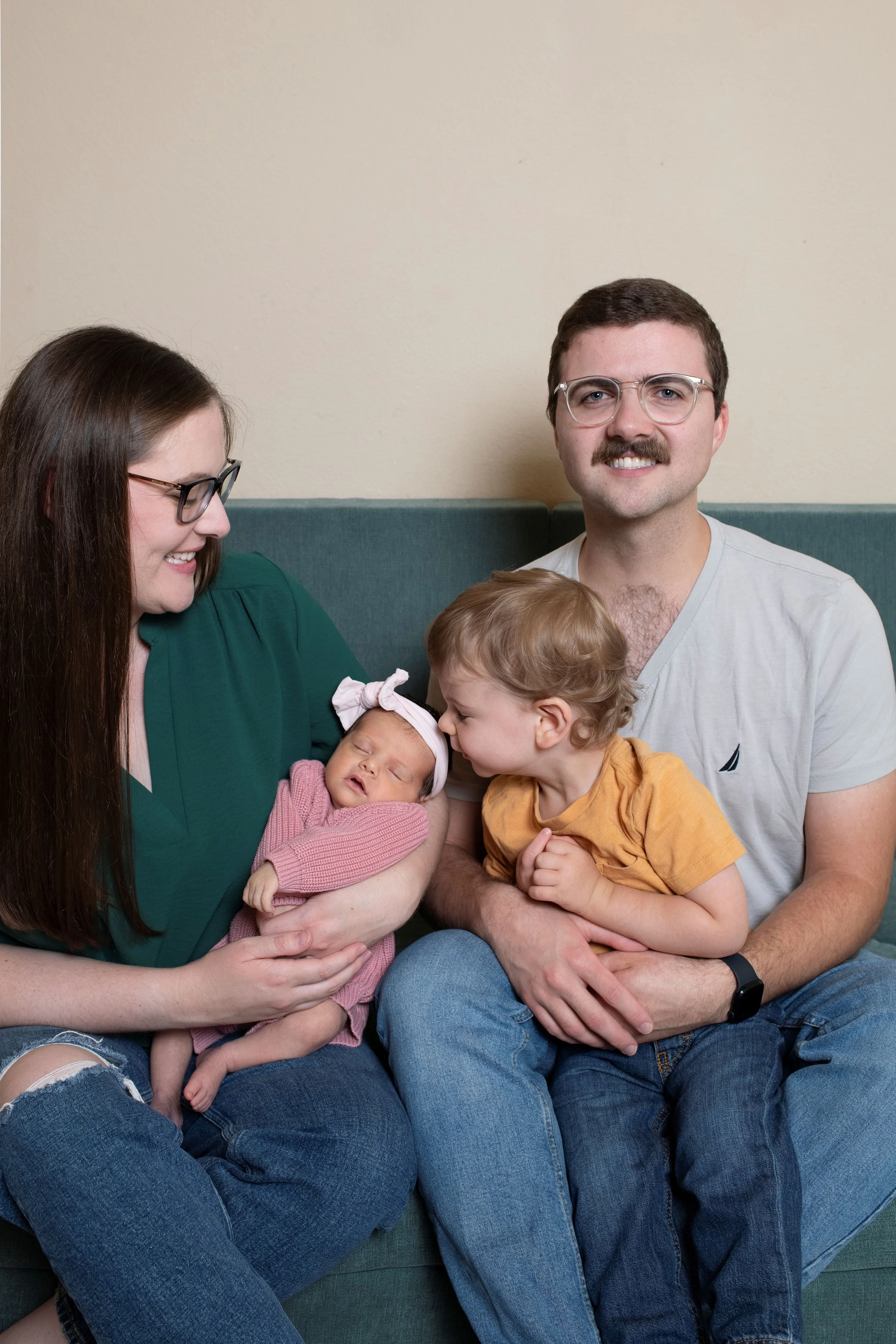A family of four sitting on a couch, including a woman holding a newborn baby, a young boy kissing the baby's face, a man smiling at the camera, all gathered in a cozy home.