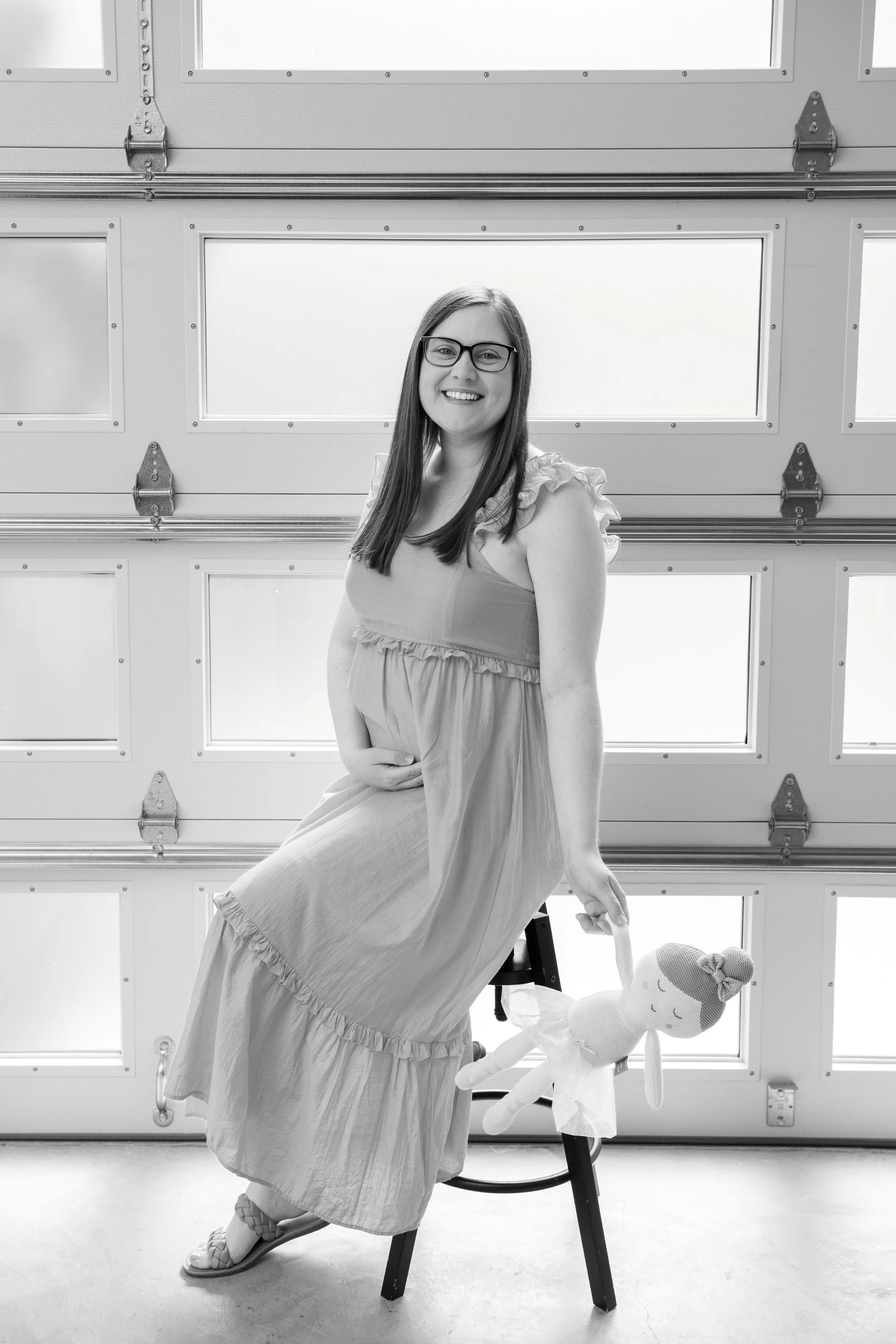 A pregnant woman sitting on a stool and holding a doll, smiling with glasses, in front of a garage door.