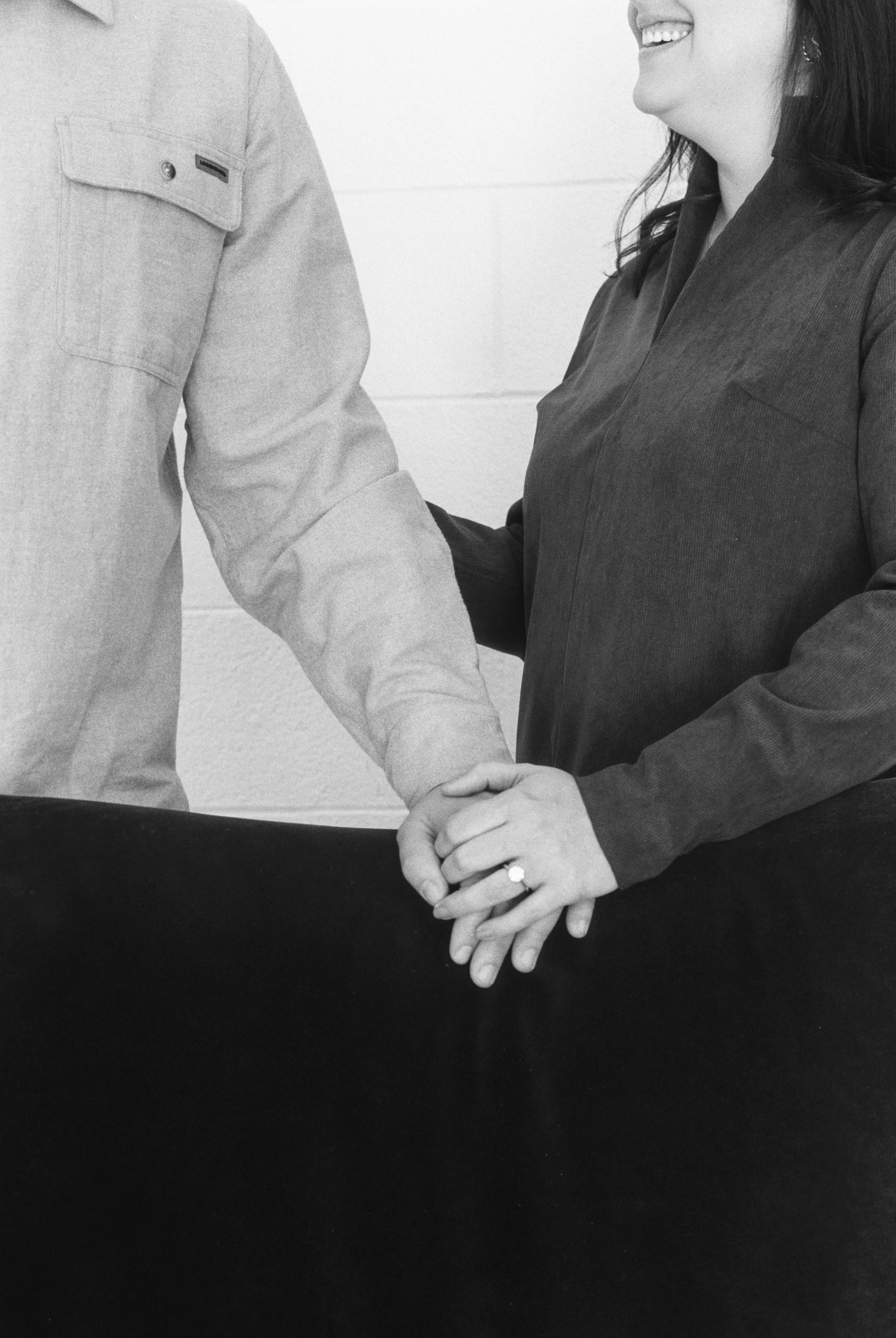 A woman with dark hair is holding hands with a man, with their hands resting on a dark surface. The woman is smiling and is wearing a ring on her left hand.