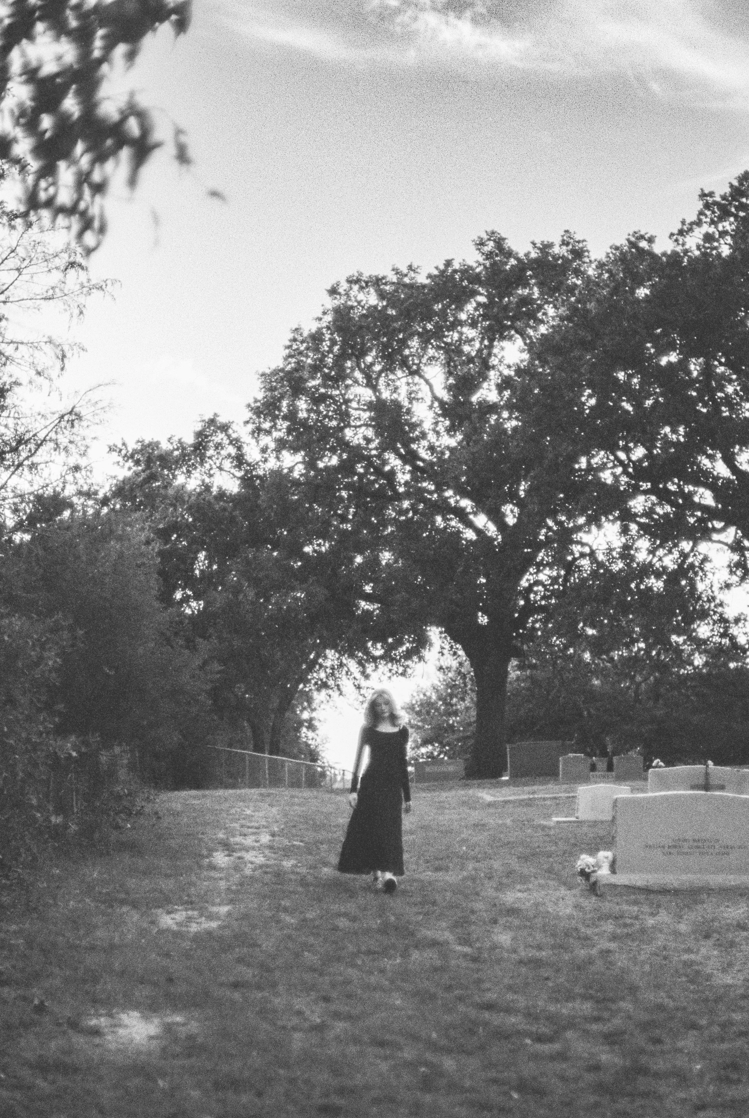 A woman in a long black dress walking alone through a cemetery with large trees and tombstones around.