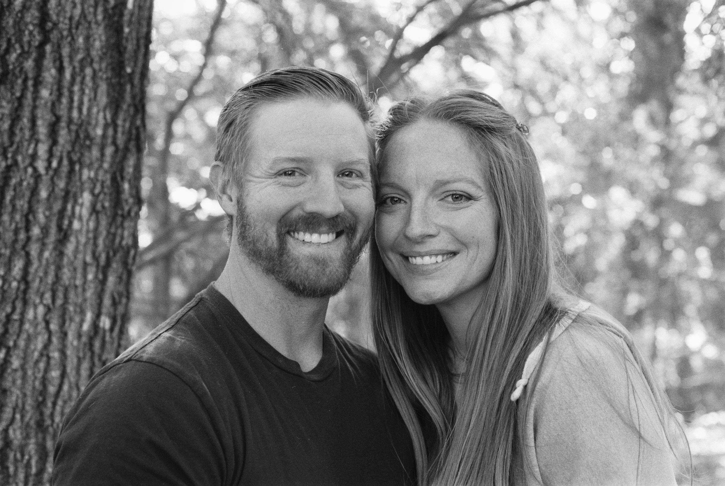A black and white photo of a smiling couple standing close together outdoors, with trees in the background.