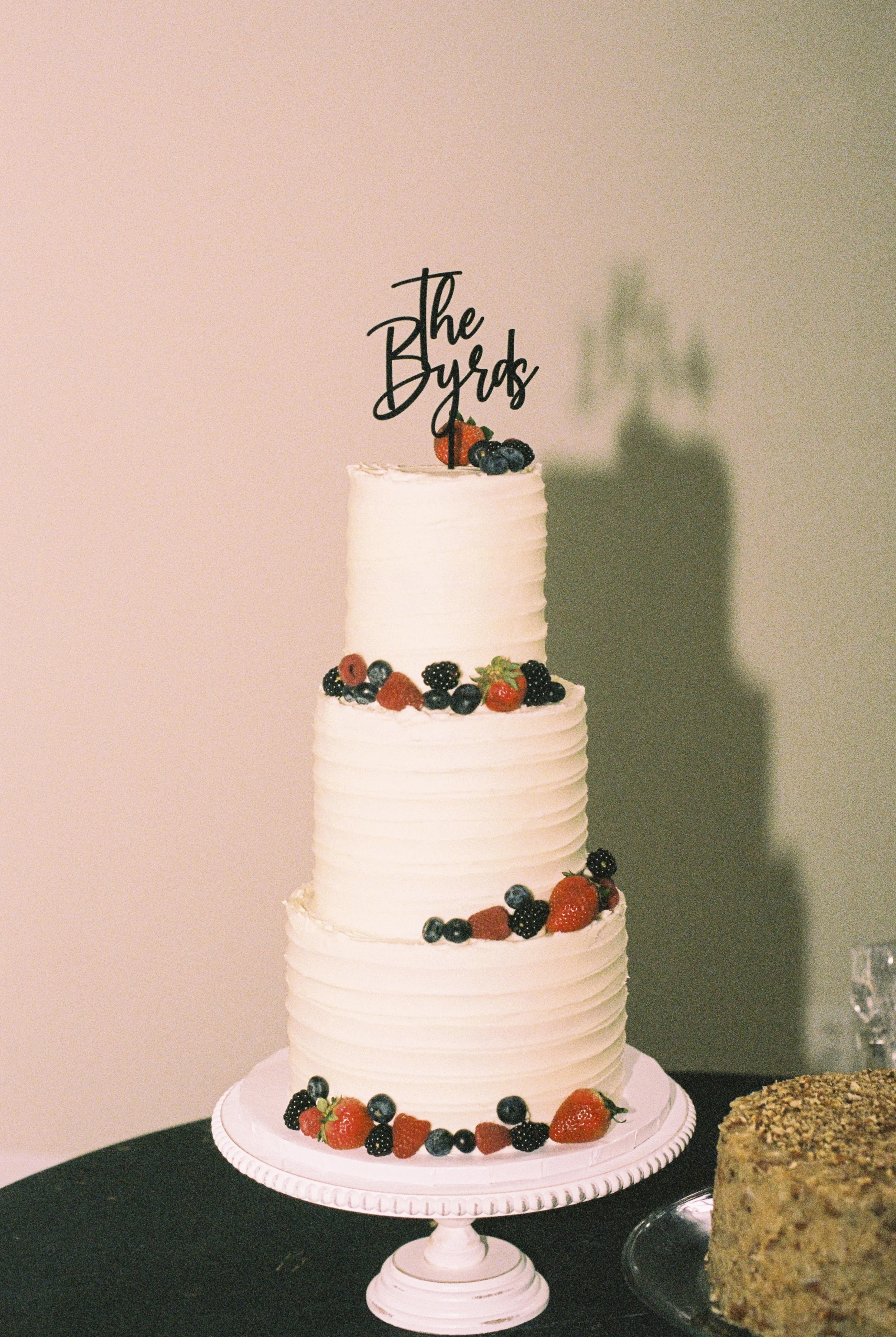 A multi-tiered white wedding cake decorated with fresh berries on each layer, topped with a black cursive cake topper that reads 'the Byrds'.