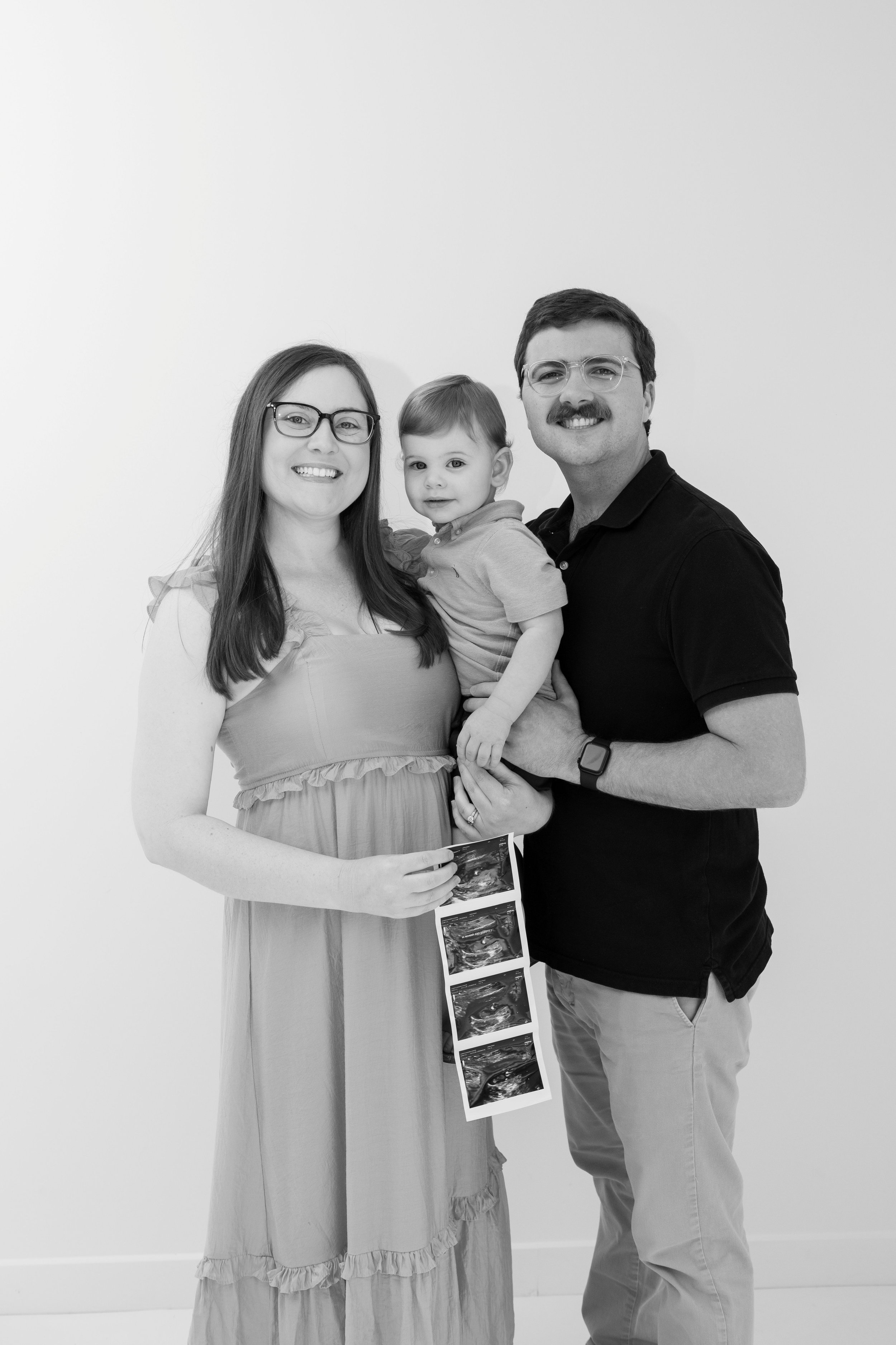 A smiling family of three, a woman, a man, and a young boy, holding ultrasound images, standing against a plain white wall.