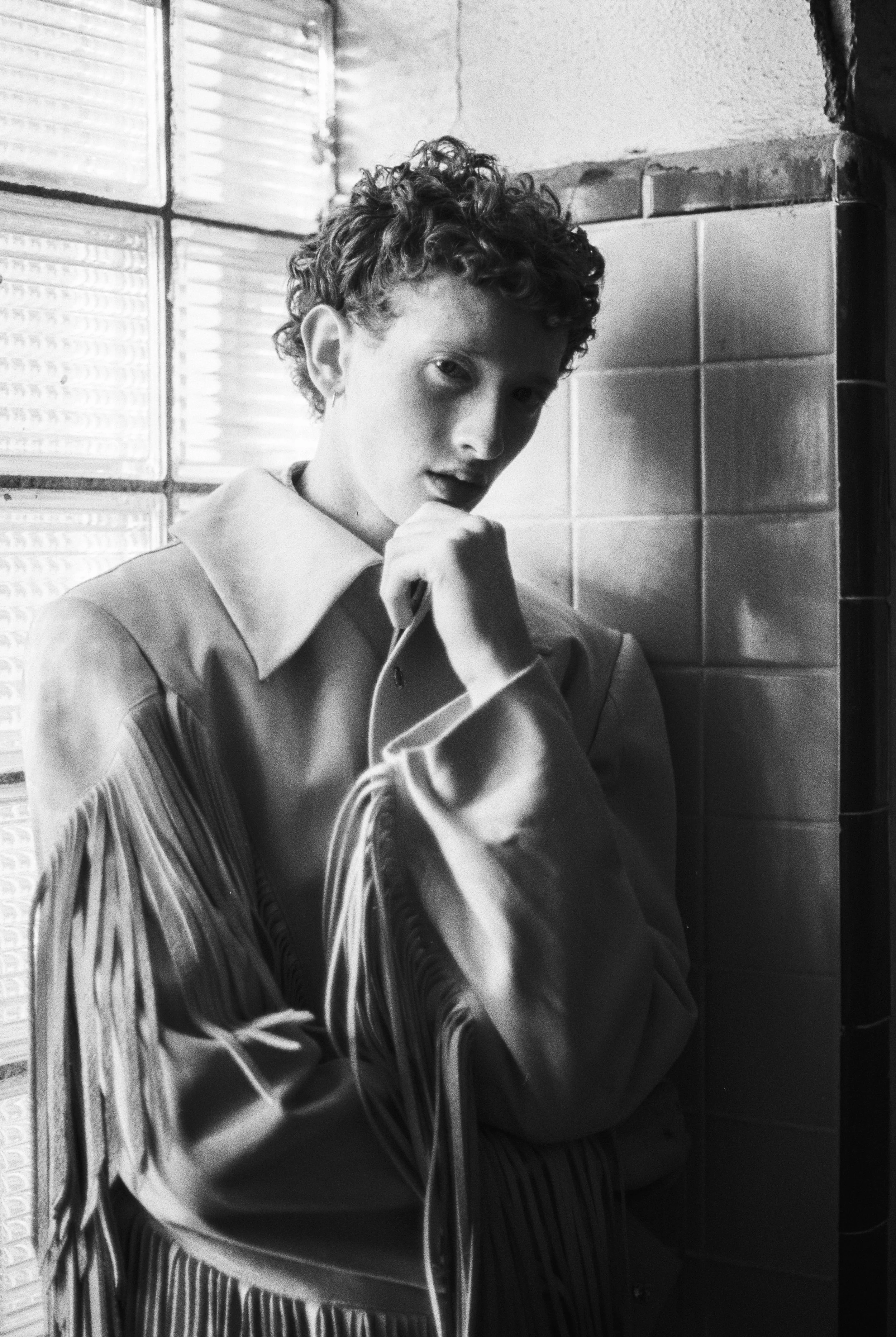 Black and white photo of a male model with short curly hair, wearing a jacket with fringe, standing against a tiled wall near a window, with her hand near her face, looking thoughtfully at the camera.