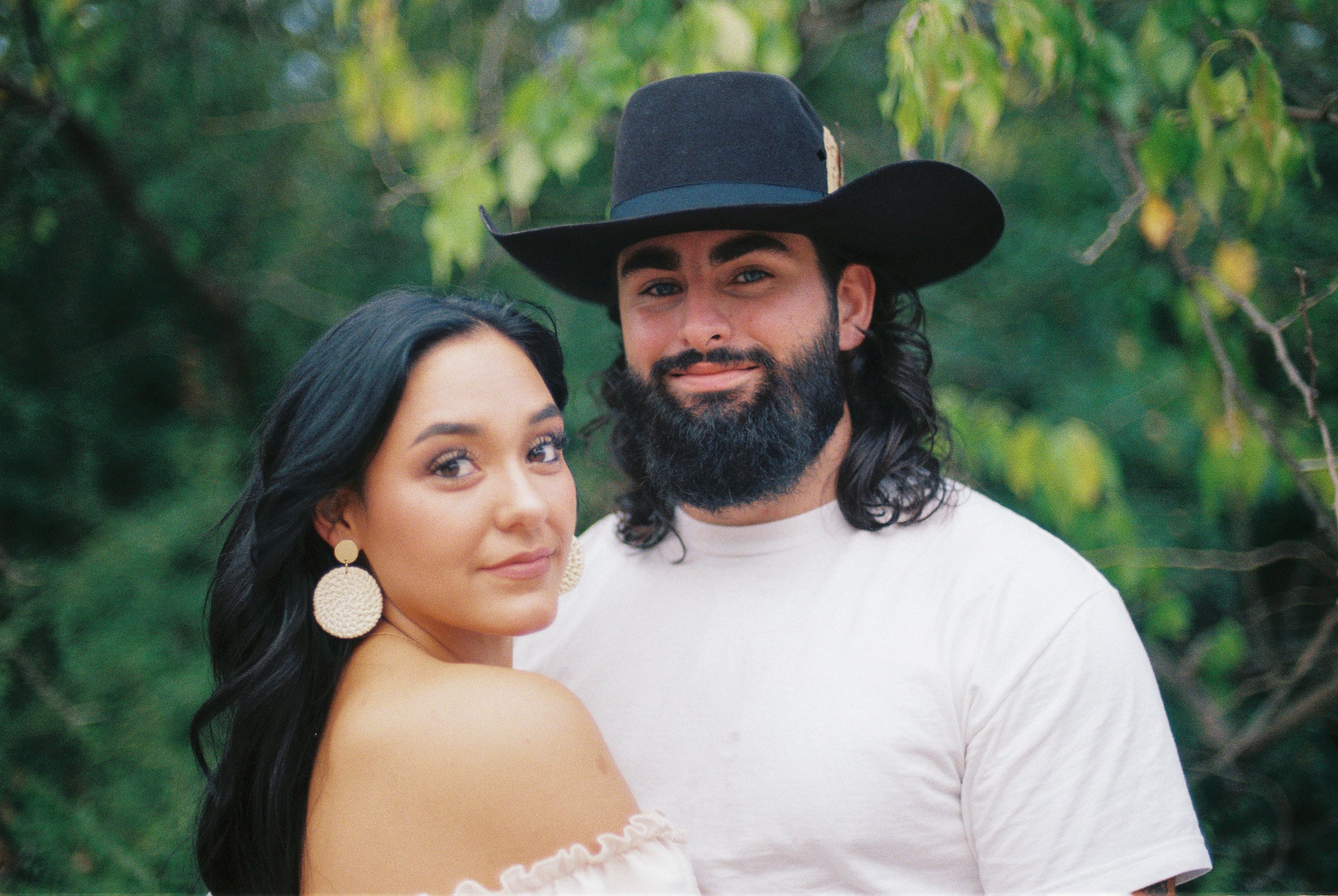 A couple standing outdoors among green trees, with the woman on the left and a man on the right wearing a black cowboy hat and white t-shirt. The woman has long dark hair, wears large round earrings, and has a slight smile, while the man has a beard,