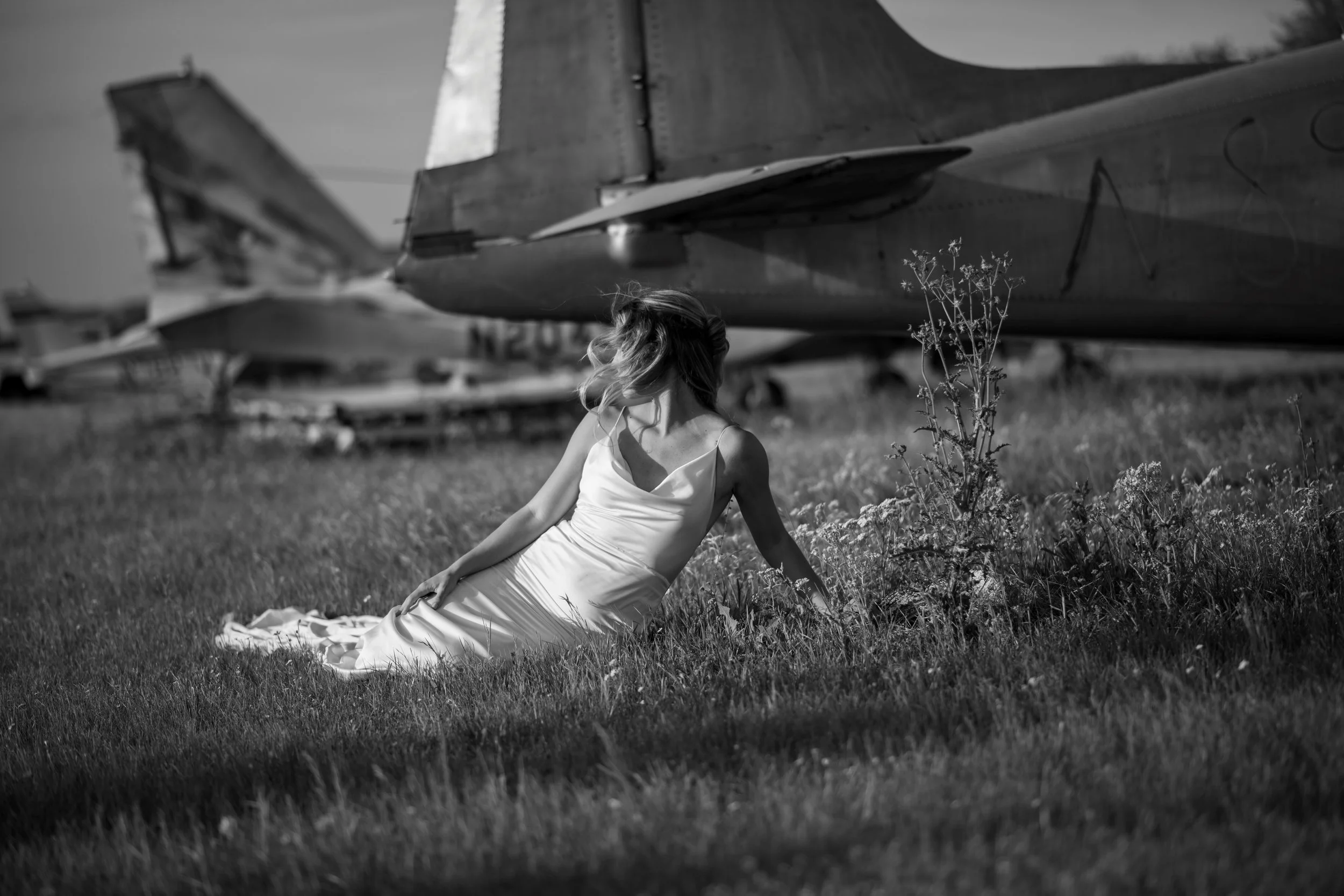 A woman in a white dress sitting on the grass in front of an airplane fuselage with her hair blowing in the wind.
