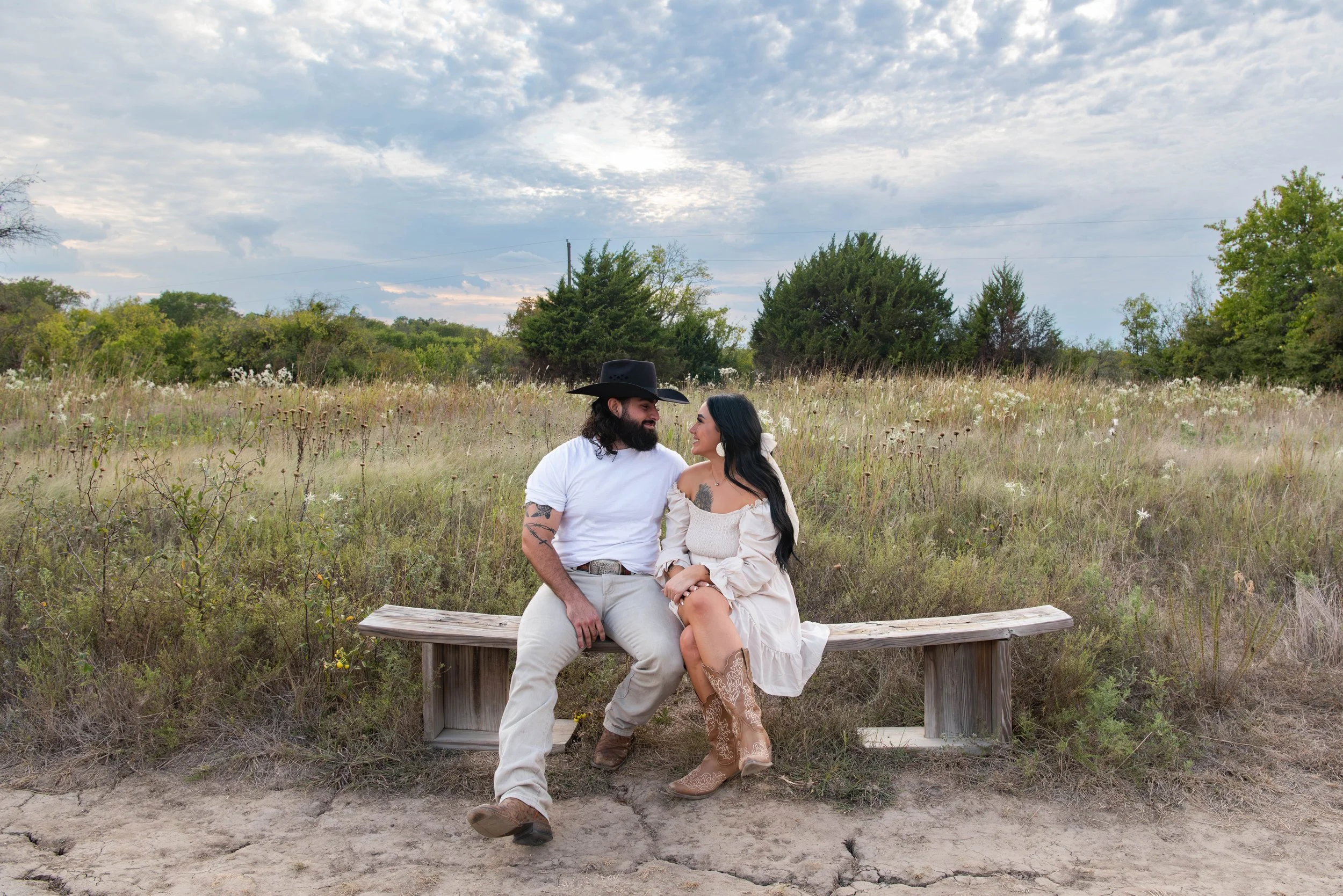 A couple sitting on a wooden bench in a field of tall grass and wildflowers, looking at each other and smiling, with trees and cloudy sky in the background.