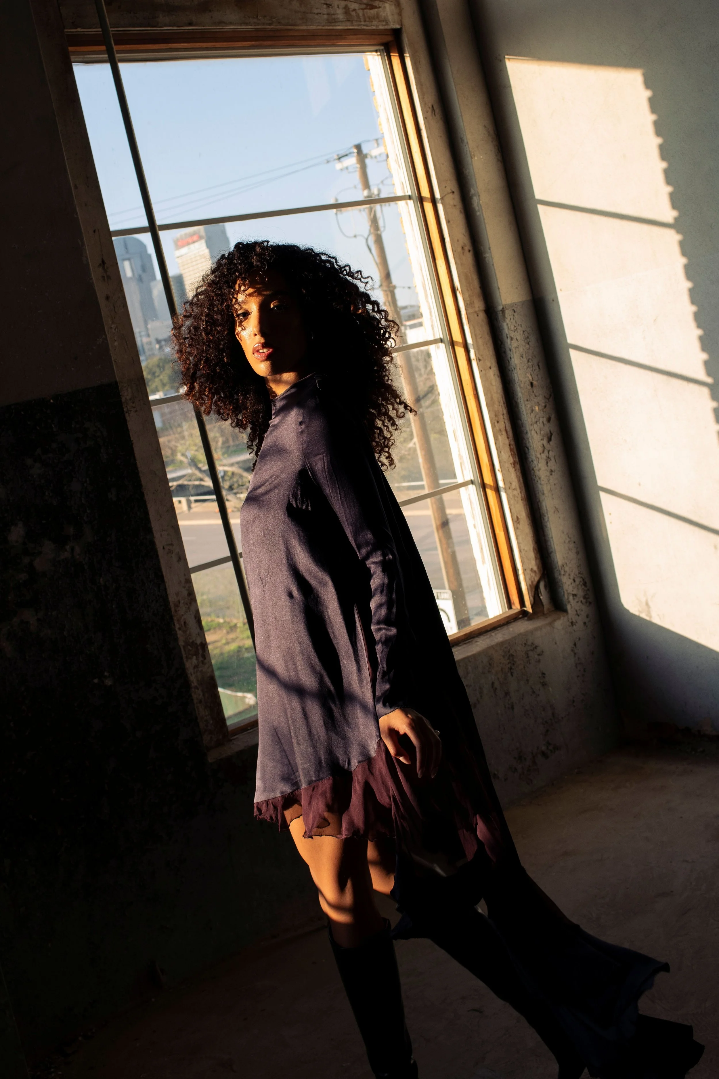 A woman with curly hair standing in front of a large window with sunlight streaming inside. She is wearing a dark jacket and a layered dress or skirt, with a cityscape visible outside through the window.