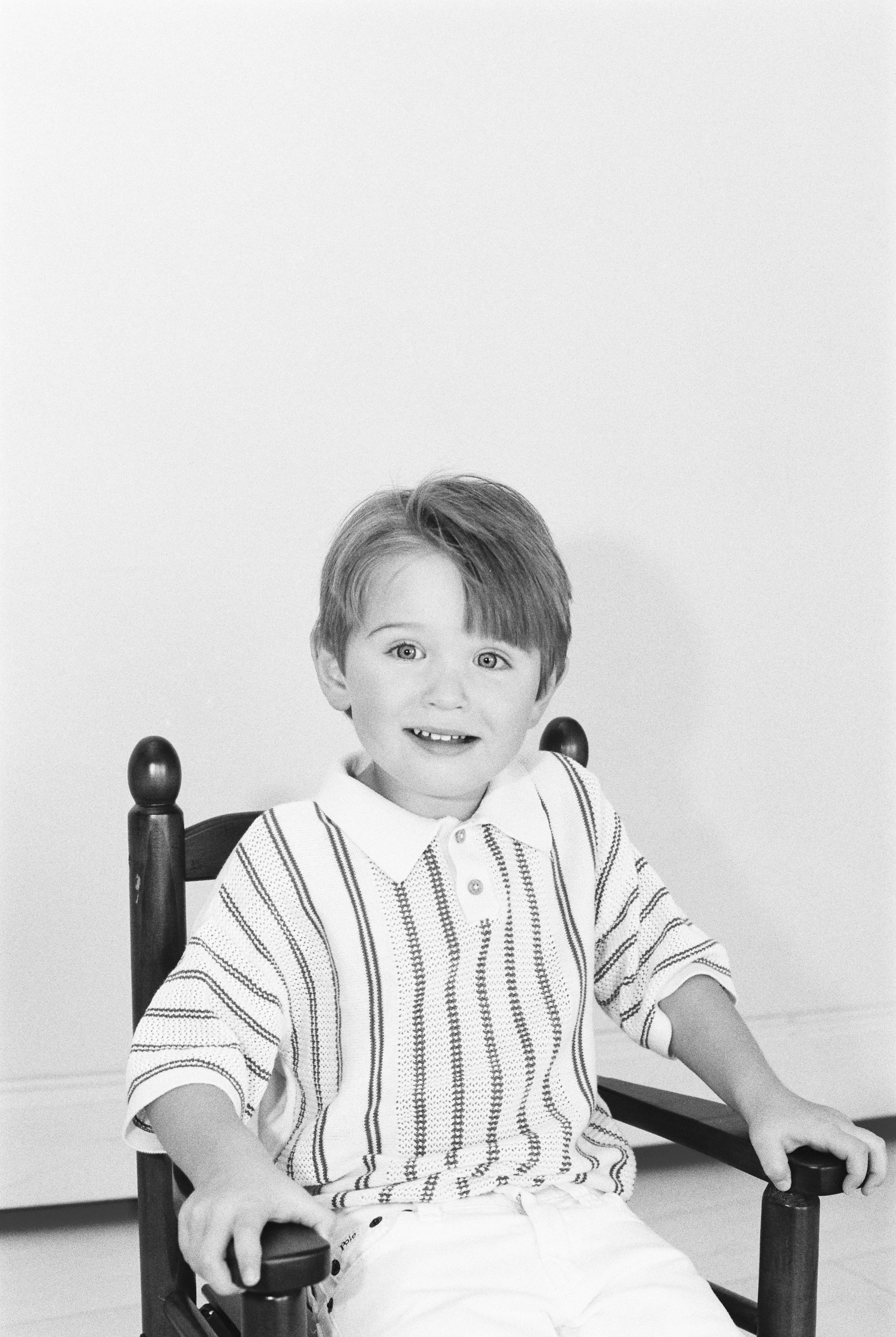 A black-and-white photo of a young boy with light hair, sitting on a wooden chair, smiling at the camera, wearing a striped polo shirt and white pants.