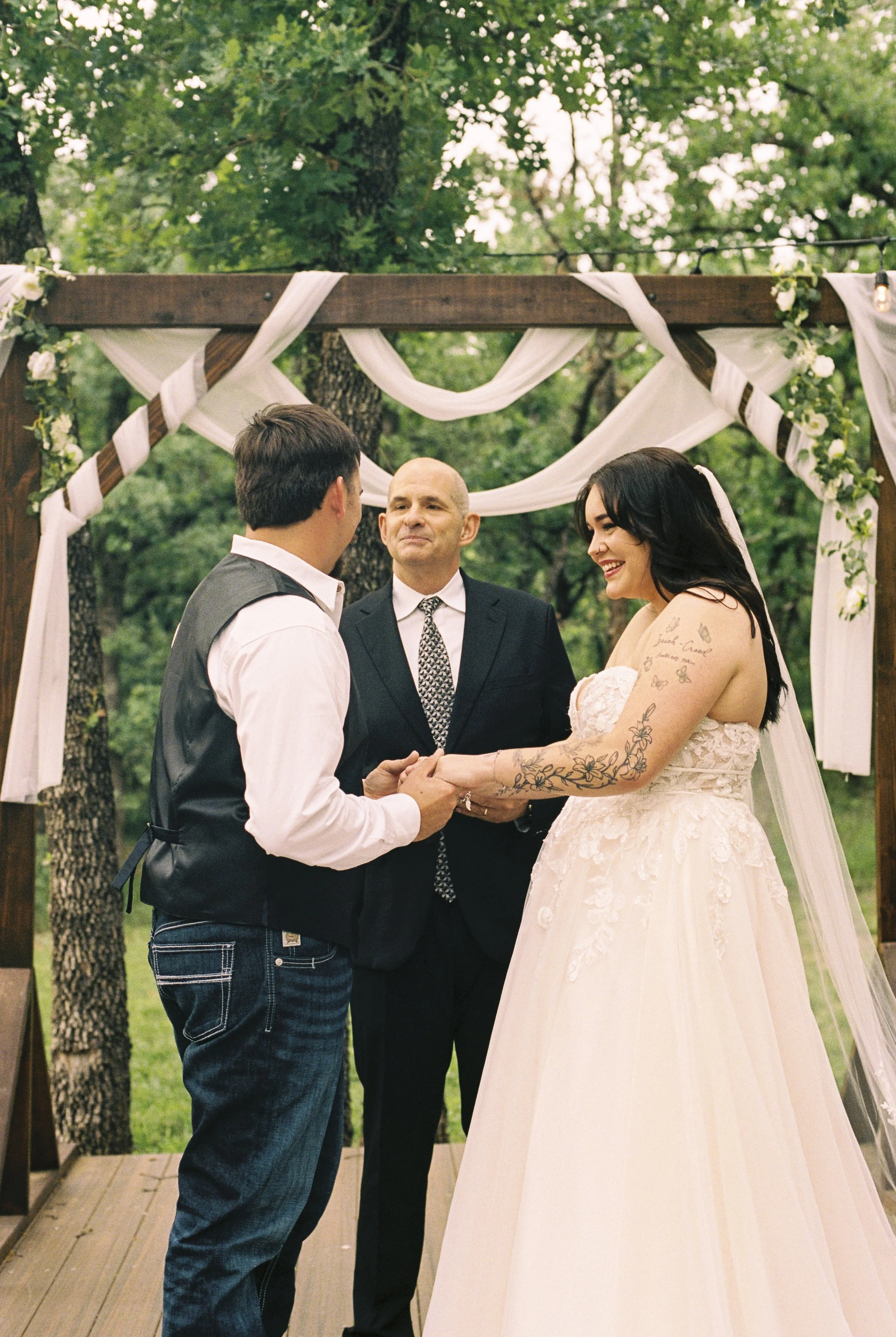 A couple getting married outdoors under a decorated arch, with a man officiating, surrounded by green trees.