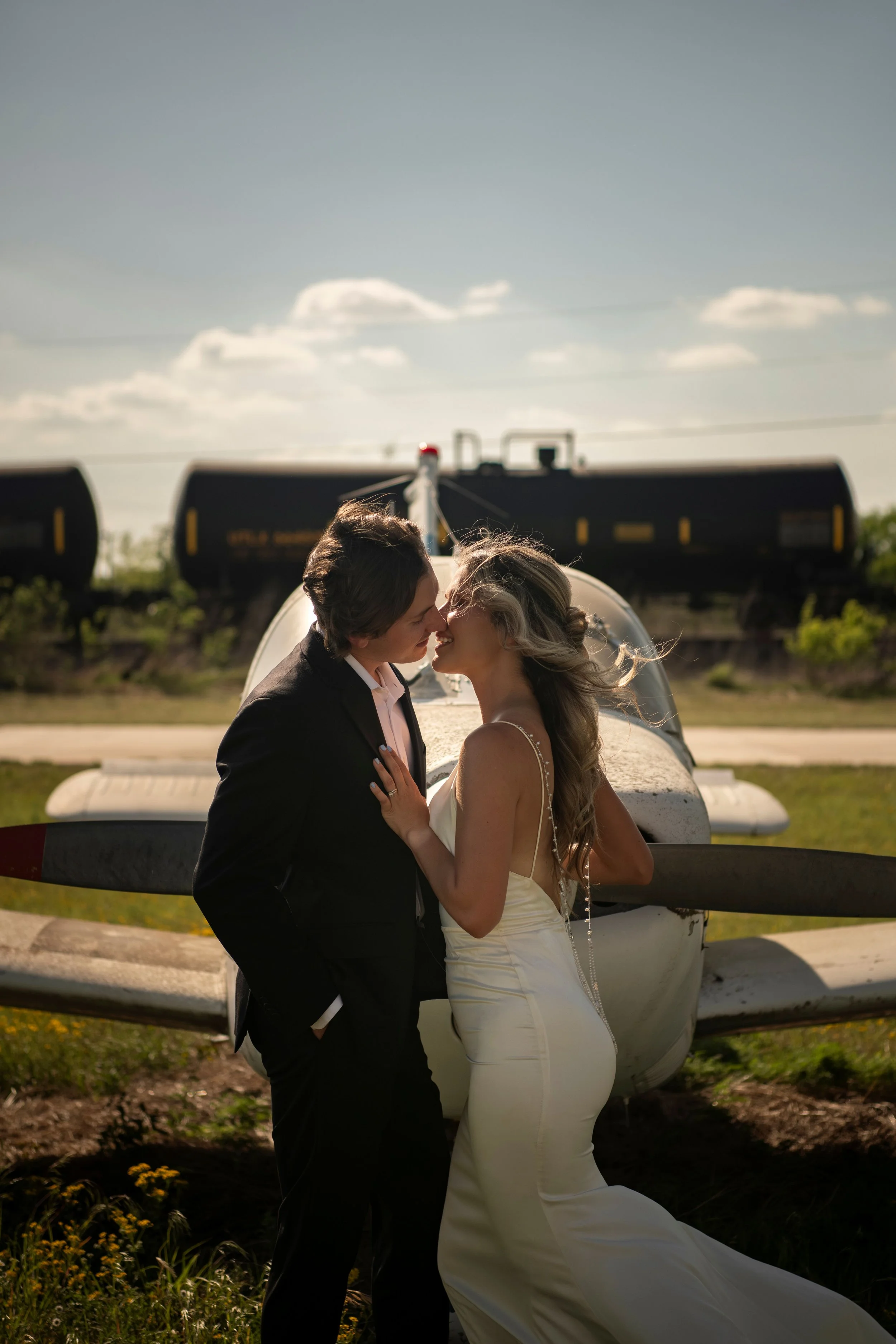 A bride and groom are in a close, intimate pose in front of an old airplane, with their noses touching and eyes closed. The bride wears a white wedding dress with spaghetti straps, and the groom wears a black suit with a white shirt. The airplane's n