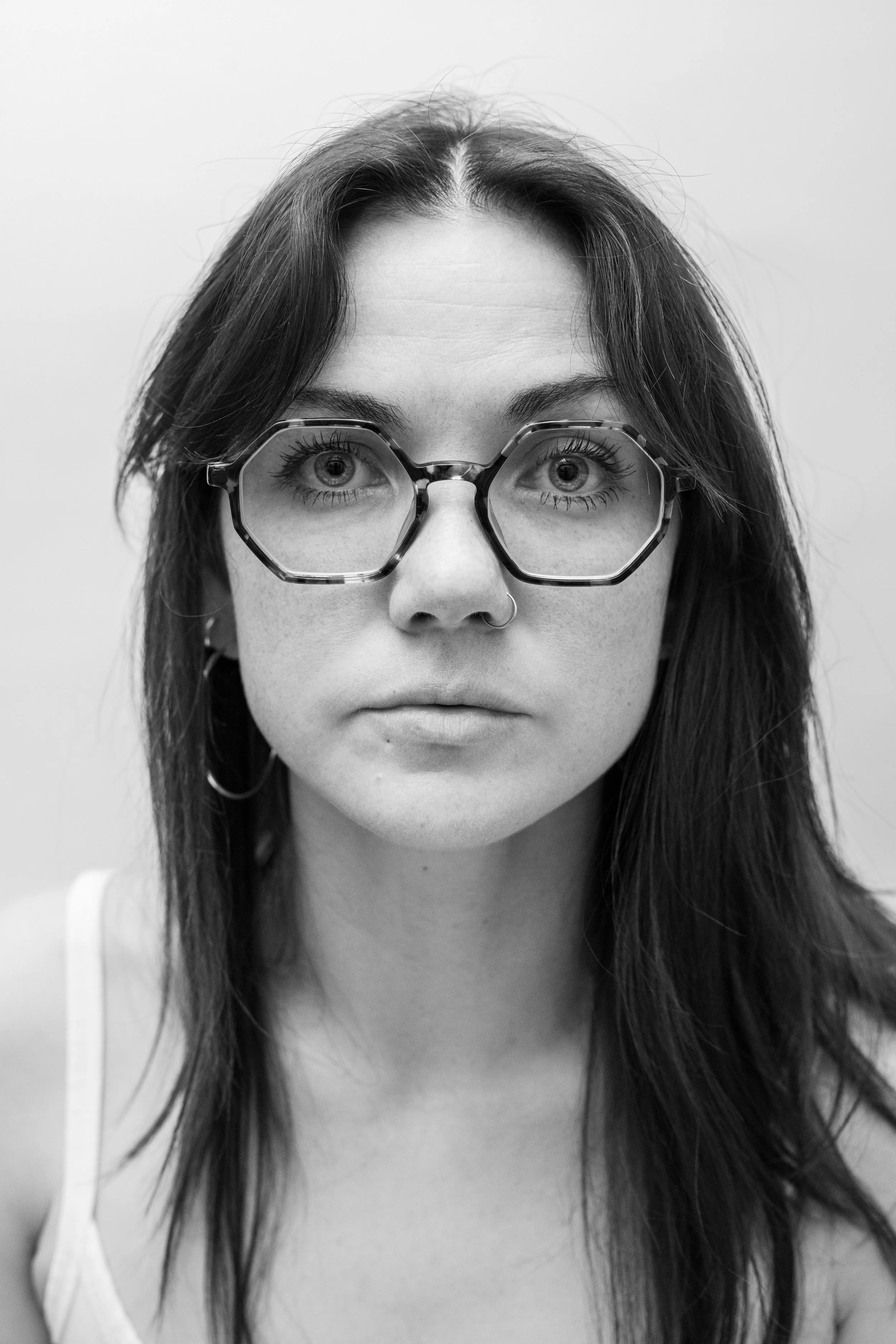 A black and white portrait of a woman with long hair, wearing large geometric glasses and hoop earrings, looking directly at the camera.