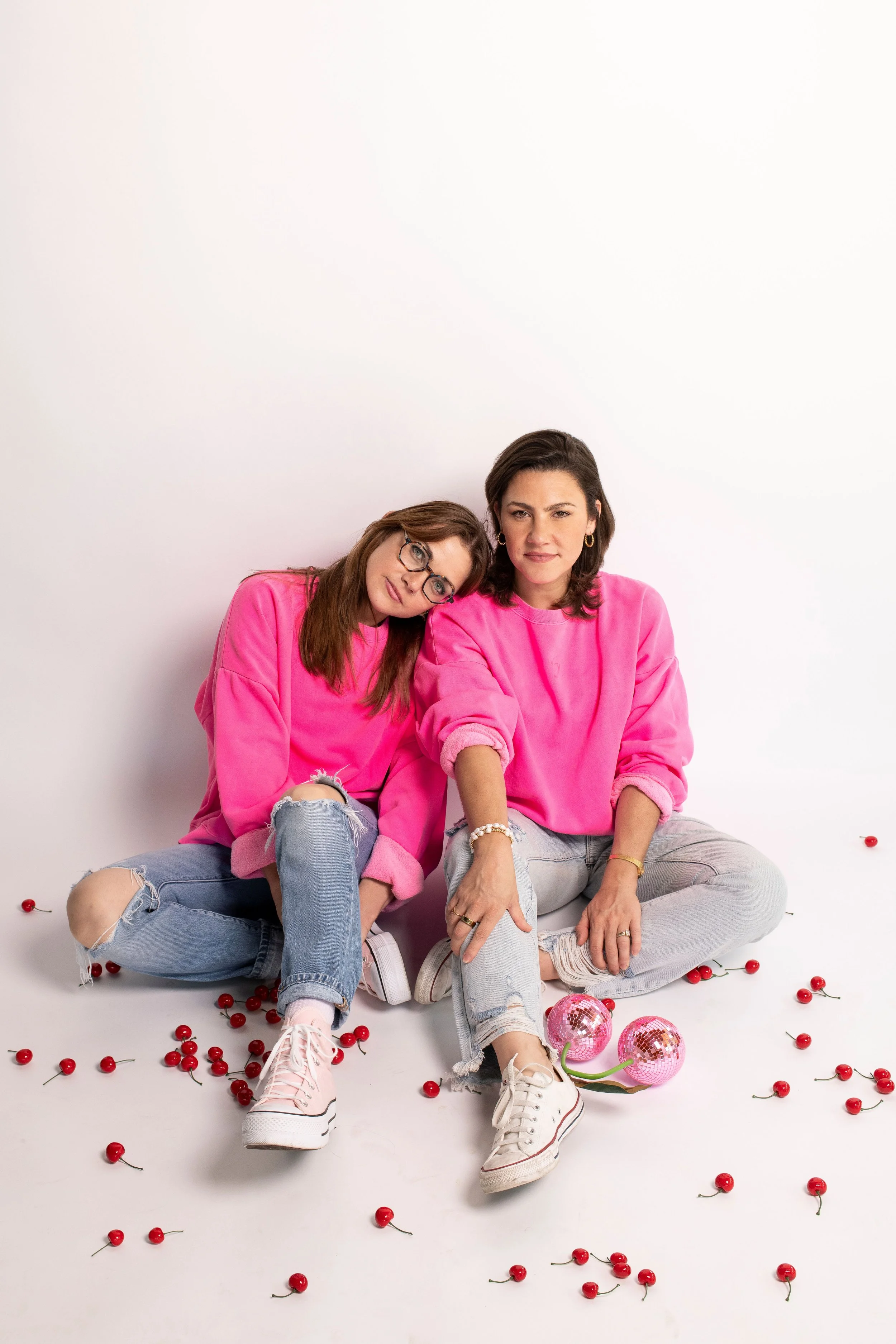Two women sitting on the floor against a plain white background, both wearing pink sweatshirts and ripped jeans. There are scattered cherries and two pink disco balls on the floor around them. One woman has glasses and is resting her head on the other's shoulder, both looking at the camera.