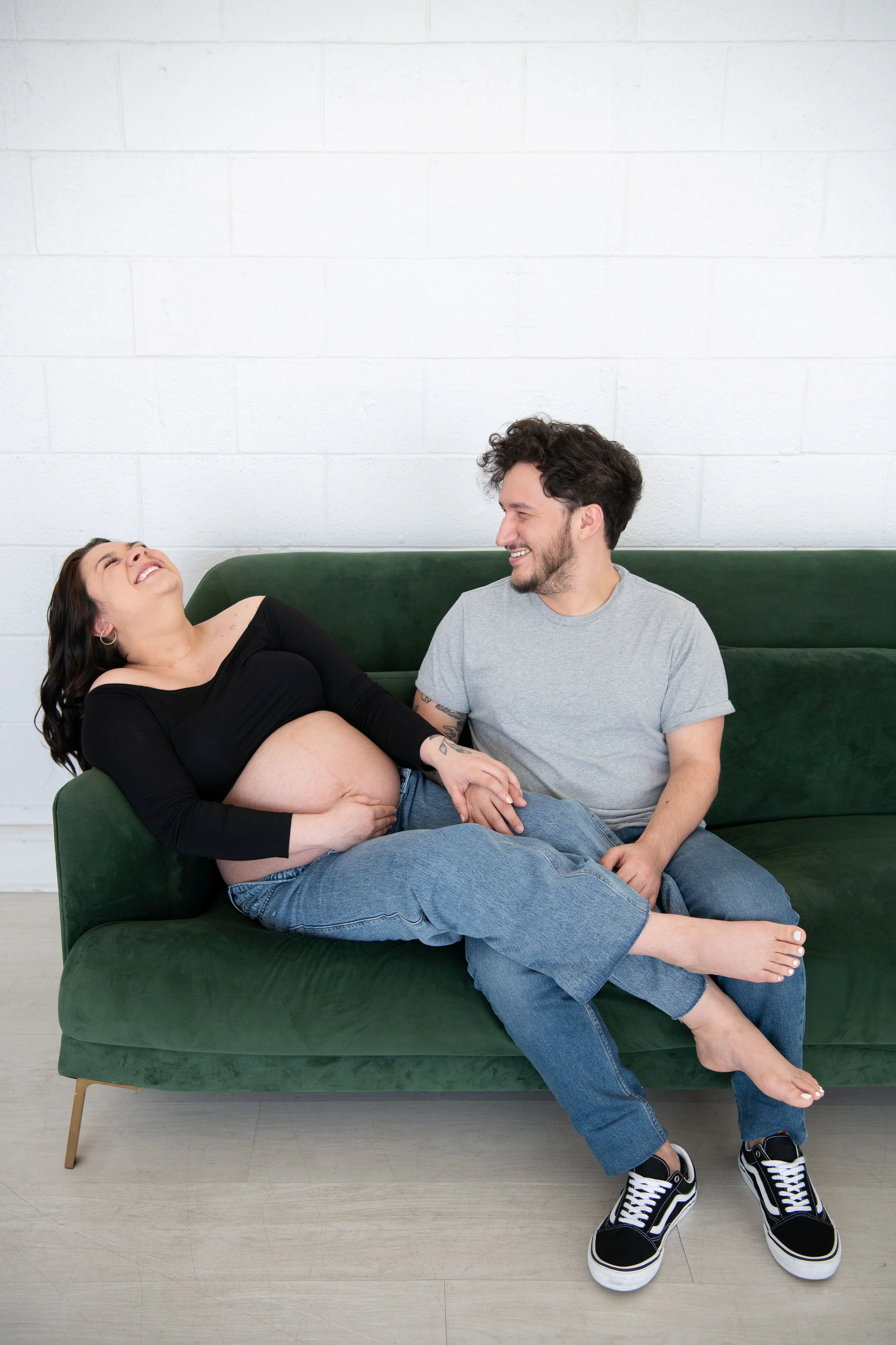 A pregnant woman and a man sitting on a green sofa, laughing and holding hands, in a modern, minimalist room.