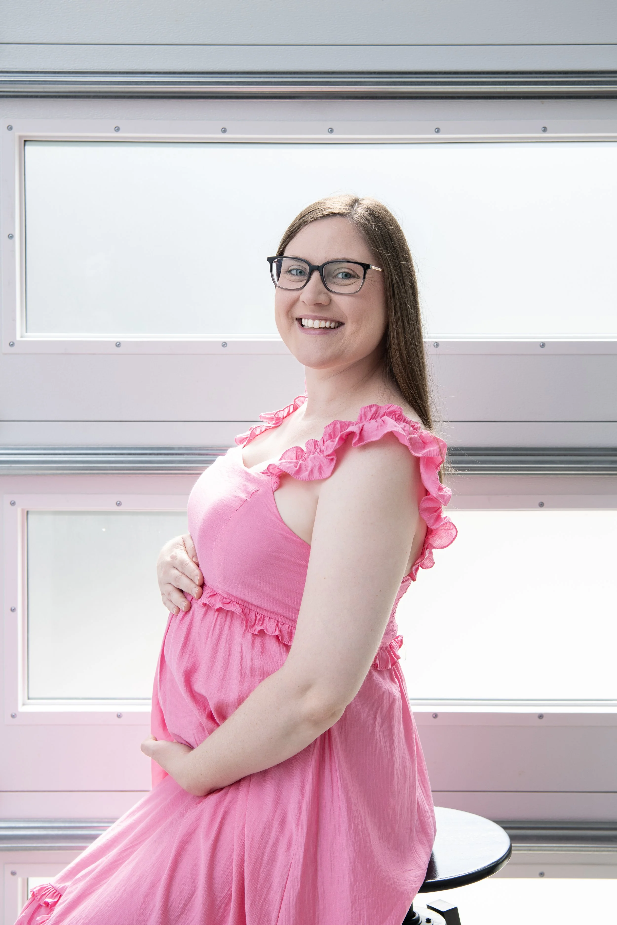 A smiling woman with glasses and long brown hair wearing a pink dress with ruffles, sitting on a stool in front of a background of large windows with frosted glass.