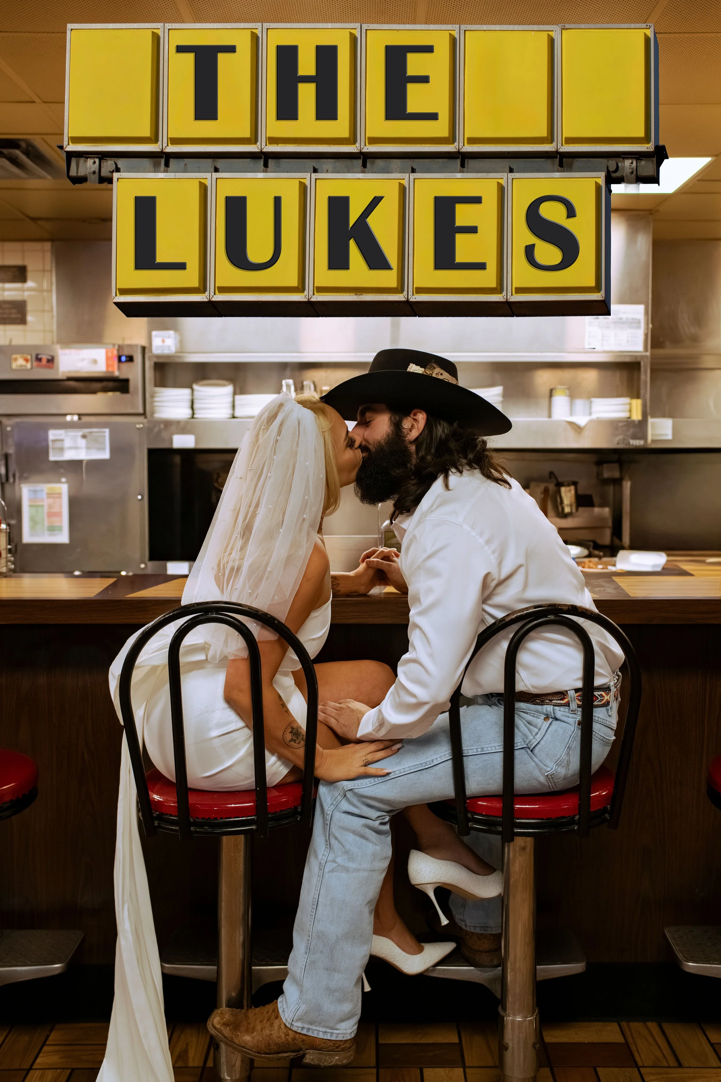 A couple is sitting on a barstool at a restaurant counter, kissing. The woman is wearing a white veil and dress, and the man is wearing a cowboy hat, white shirt, and jeans. Above them is a yellow sign that says 'The Lukes'.