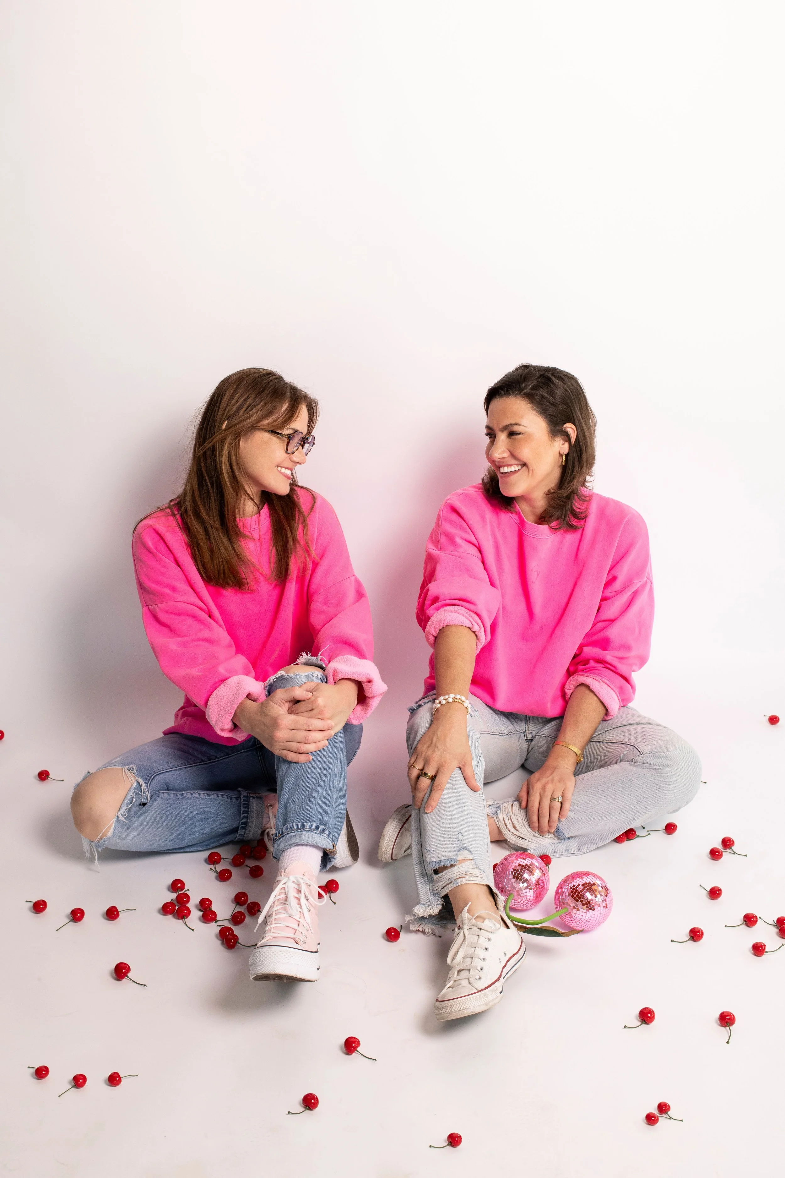 Two women sitting on the floor in pink sweatshirts and ripped jeans, smiling and looking at each other. Scattered cherries and pink disco balls around them, with one woman having a bracelet and ring, the other wearing glasses.