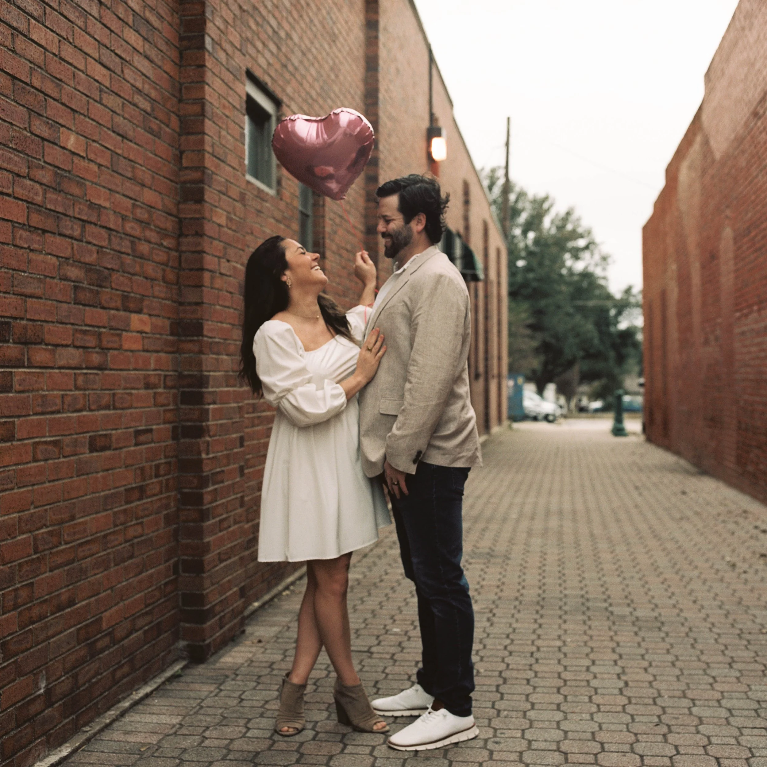 A woman in a white dress holding a pink heart-shaped balloon, smiling at a man in a beige blazer, standing in a brick alleyway.