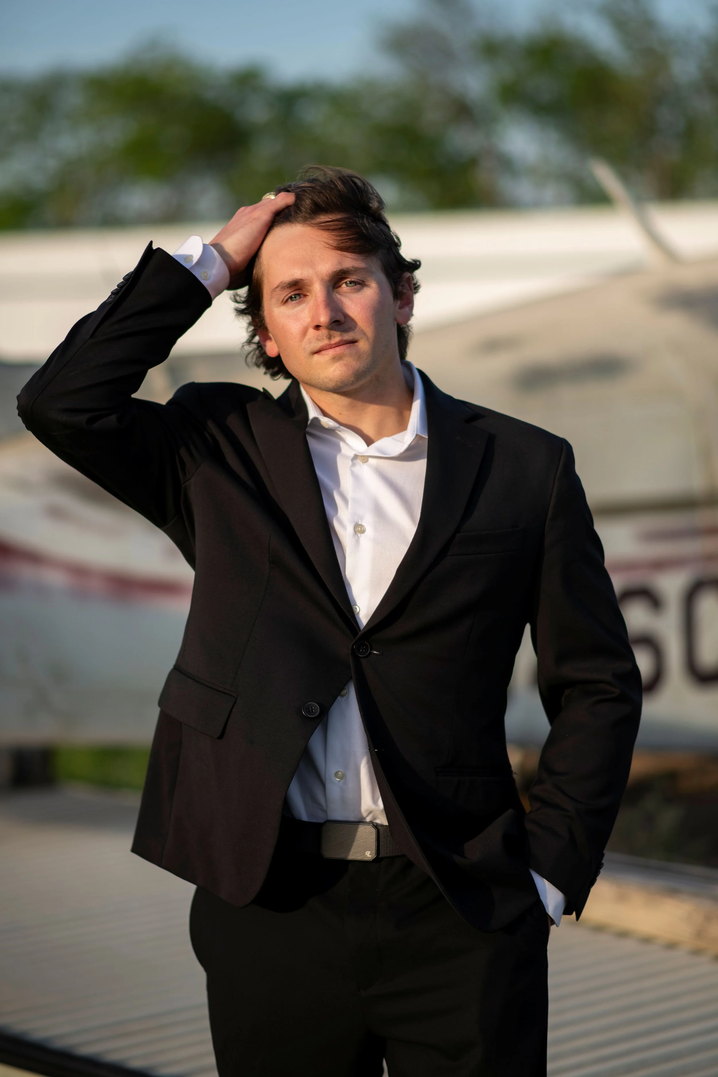 A young man in a black suit with a white shirt, standing outdoors with a blurred background of trees and a boat, touching his hair with a serious expression.