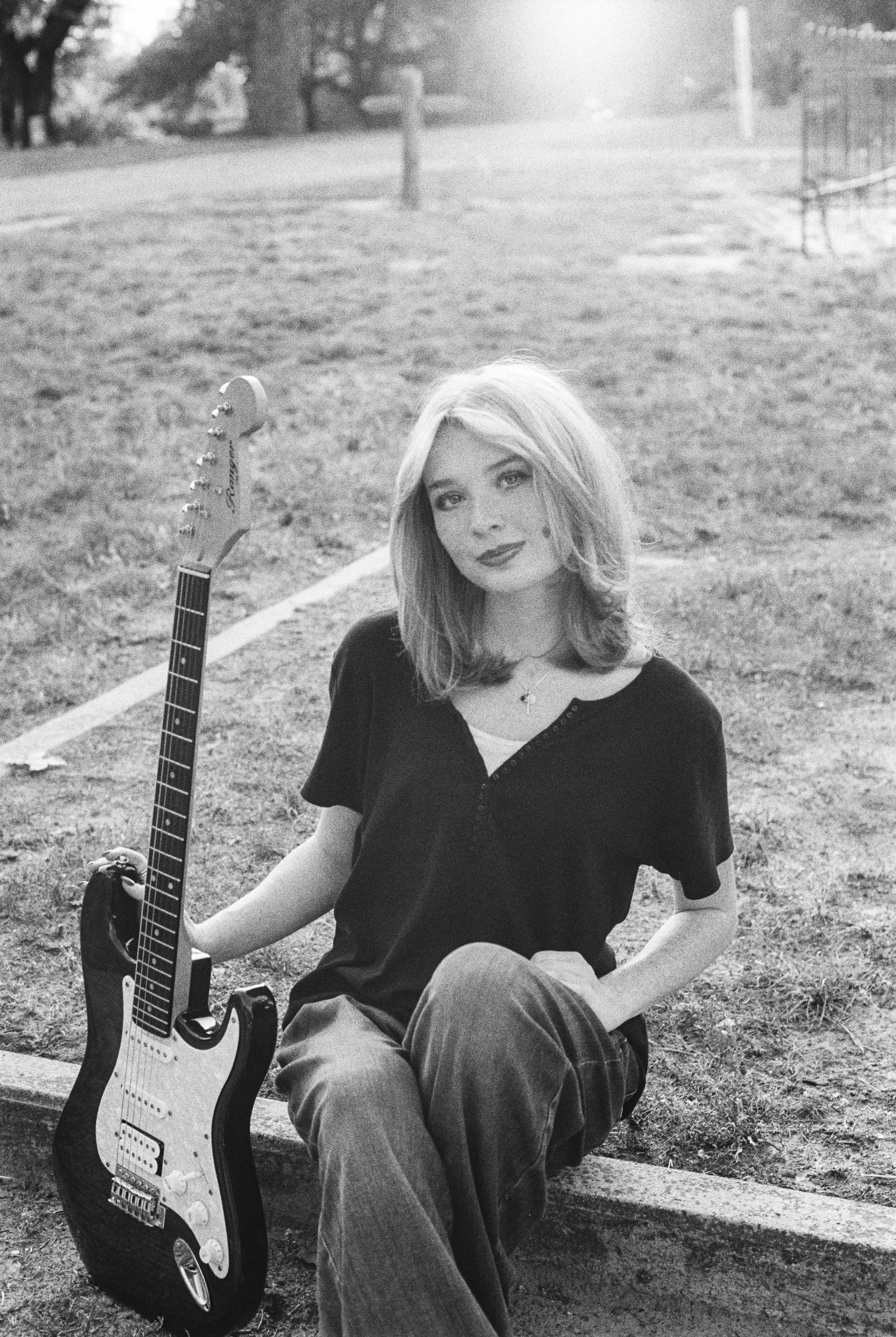 A young woman with shoulder-length hair sitting outdoors on a concrete ledge, holding an electric guitar. She is wearing a dark short-sleeve shirt and loose pants, with a necklace. The background shows a grassy area with trees and a blurred fence.