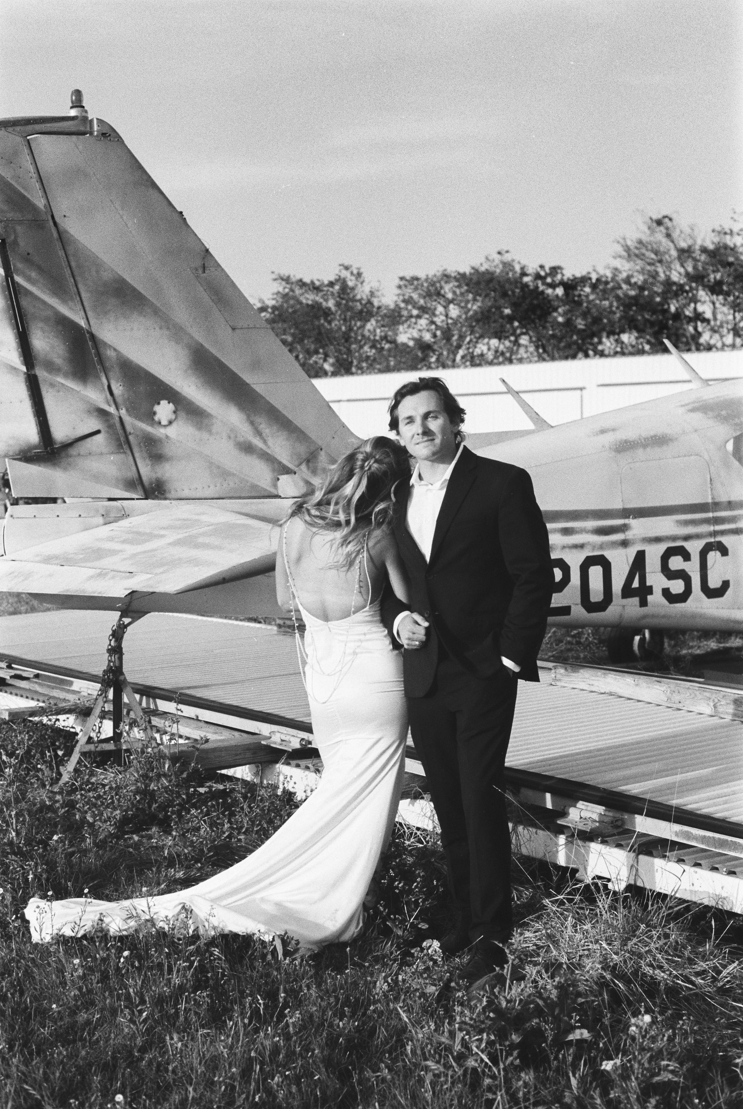 A black and white photo of a bride and groom standing together in front of a small airplane. The bride wears a wedding dress and the groom wears a tuxedo. The airplane has the number 204SC on its side and is parked on a grassy area.