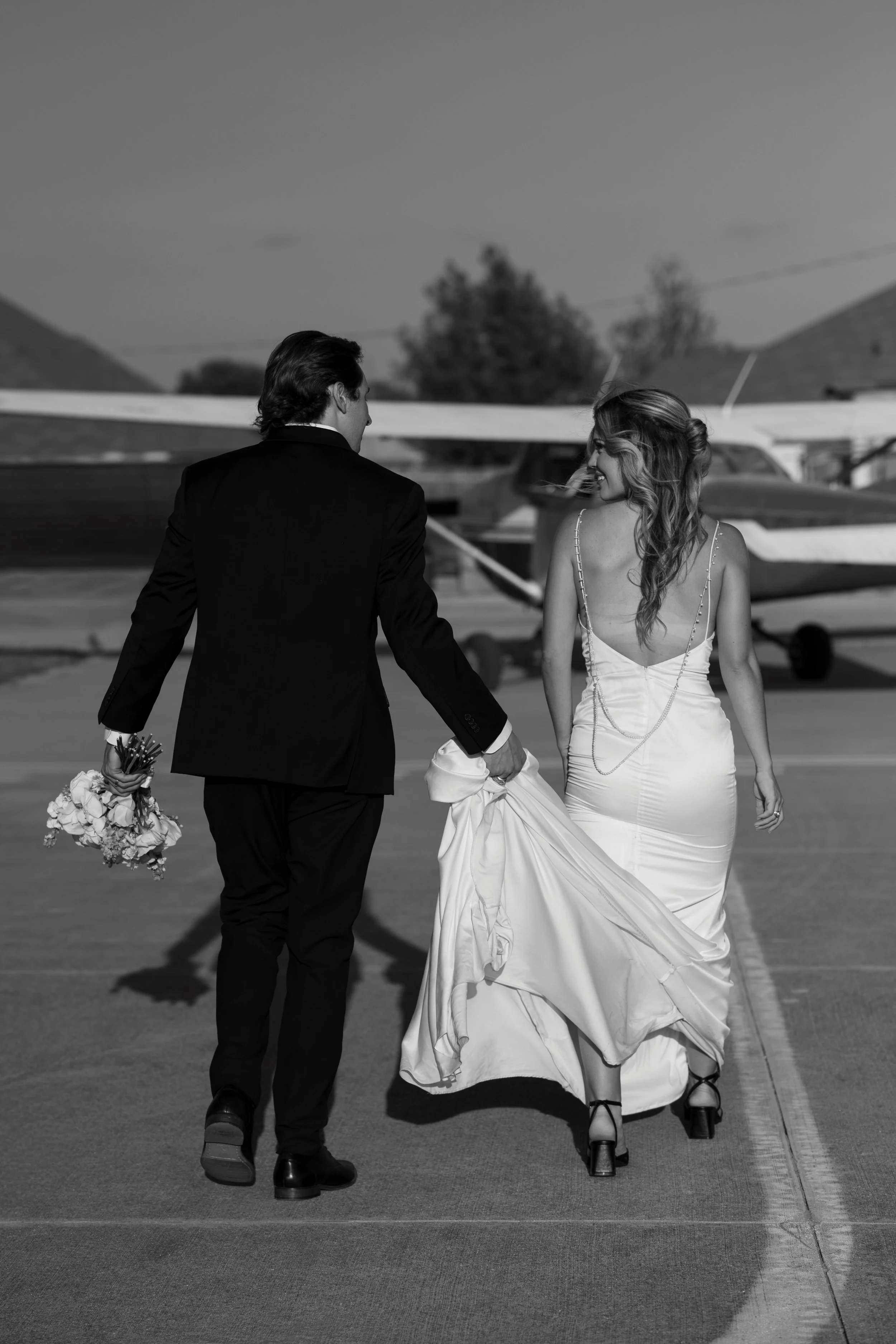 A black-and-white photo of a man and a woman dressed in wedding attire, walking hand-in-hand on an airport tarmac, with small airplanes in the background.