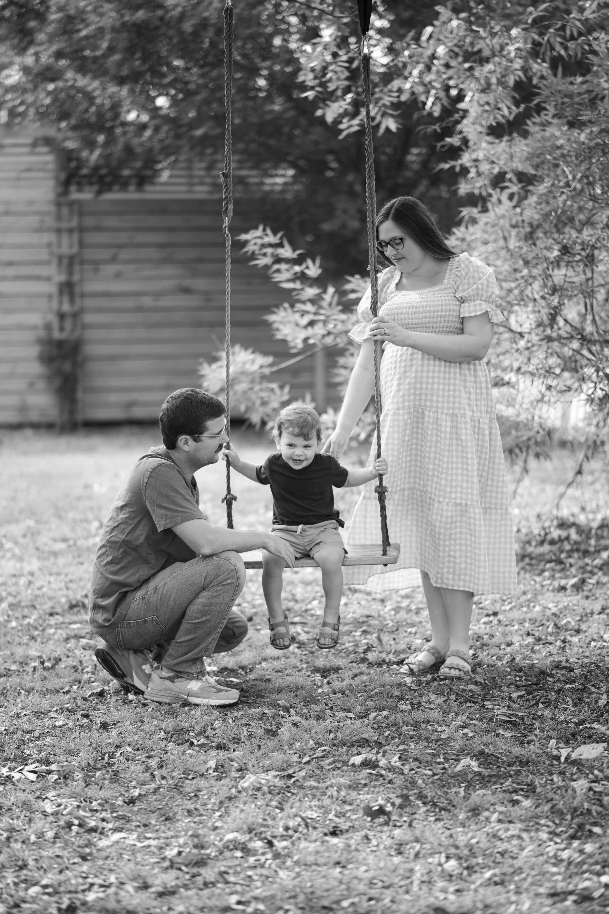 A black and white photo of a family outside with a young girl sitting on a swing, a man crouching next to her, and a woman standing holding the swing's ropes.