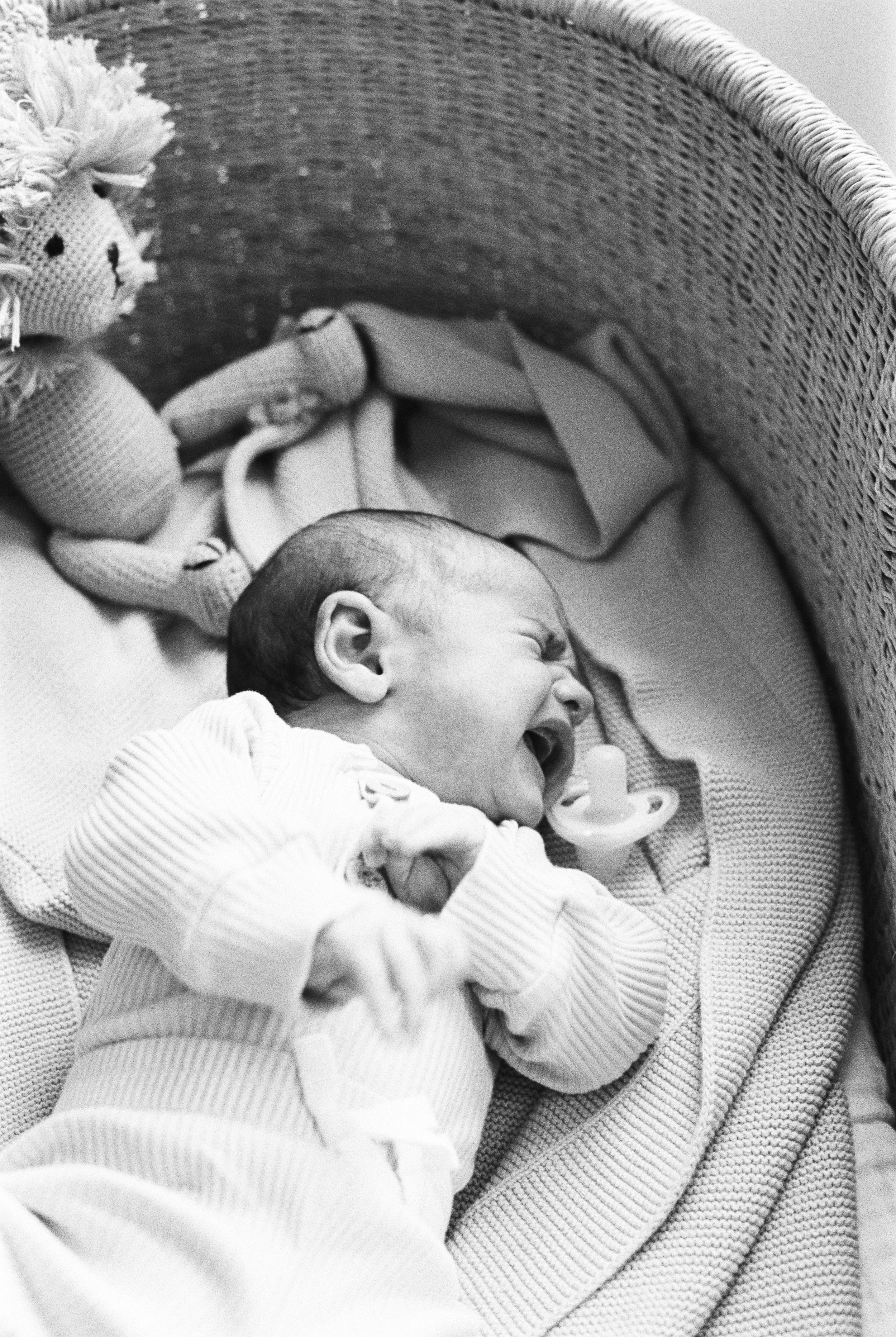 A baby lying in a crib, laughing with a pacifier in its mouth, wearing a striped onesie, and holding a toy.