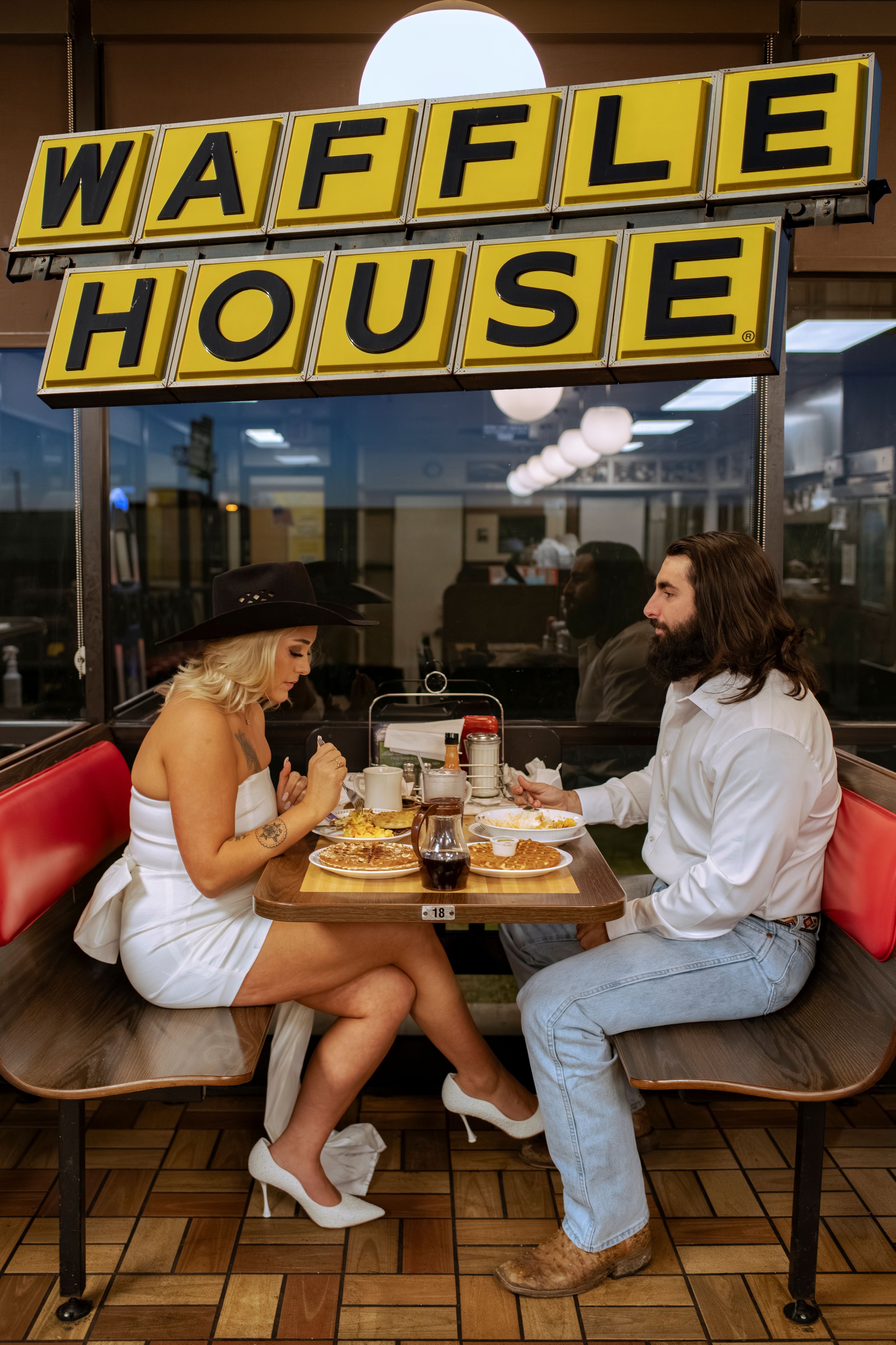 A woman and a man sit across from each other at a diner table, enjoying breakfast with waffles and coffee. The woman is wearing a white dress, white high heels, and a black cowboy hat, while the man is dressed in a white shirt and jeans, wearing cowb