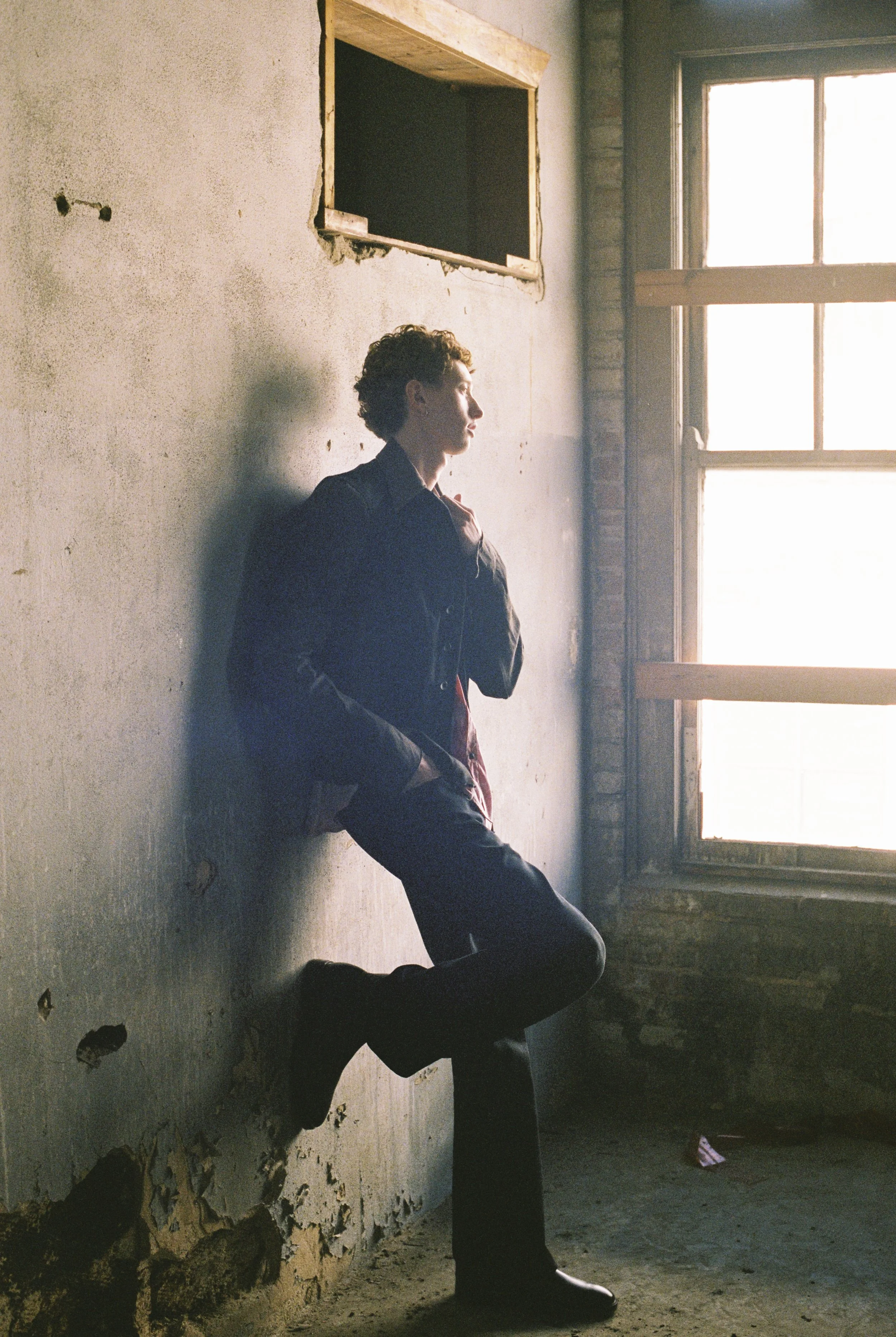 A person with curly hair, dressed in dark clothing, is kneeling against a weathered, peeling wall next to a window with natural light streaming in.