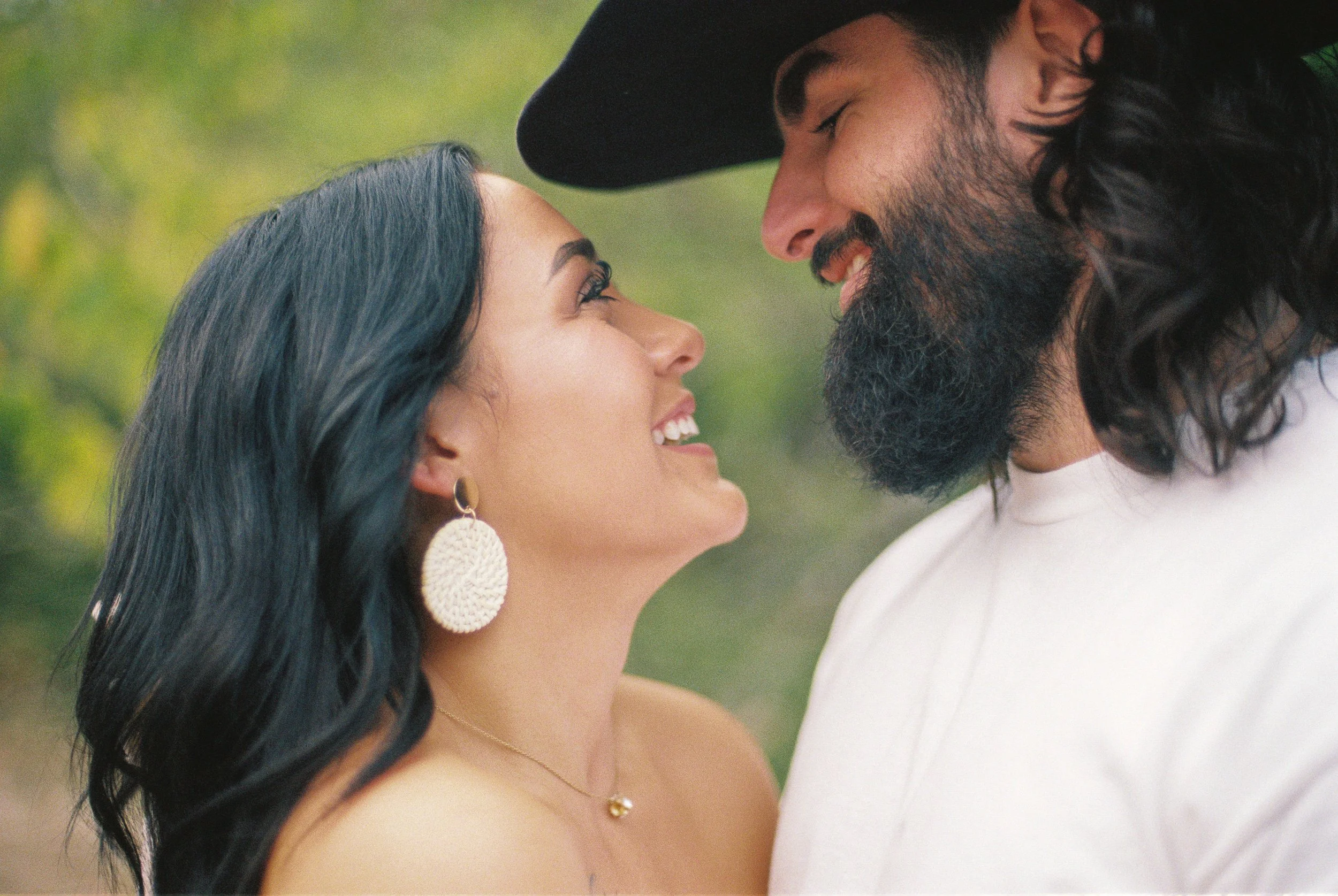 A woman and a man face to face, smiling and gazing into each other's eyes outdoors with blurred green foliage in the background.
