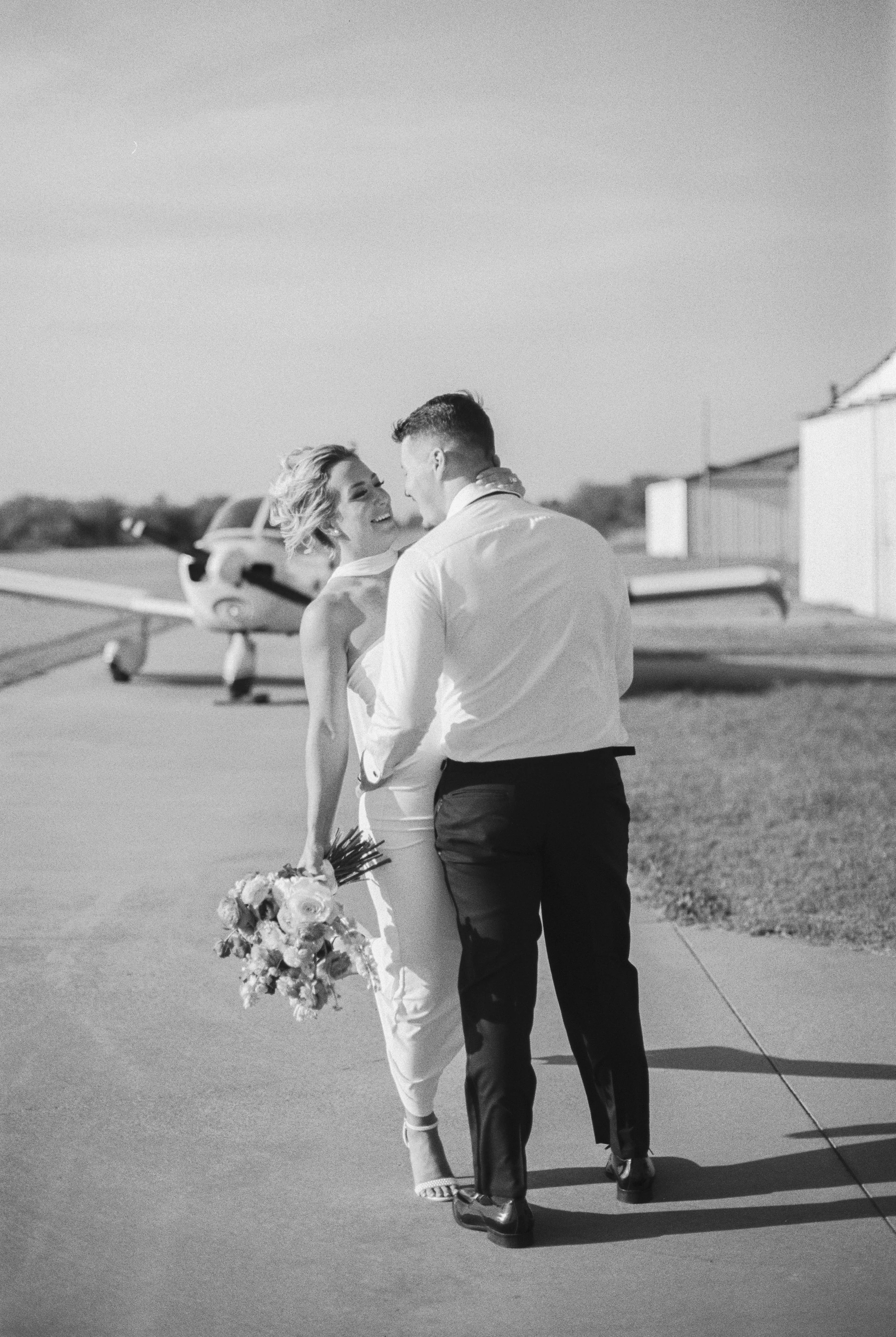 A black and white photo of a bride and groom smiling and holding each other outdoors, with small aircraft in the background.