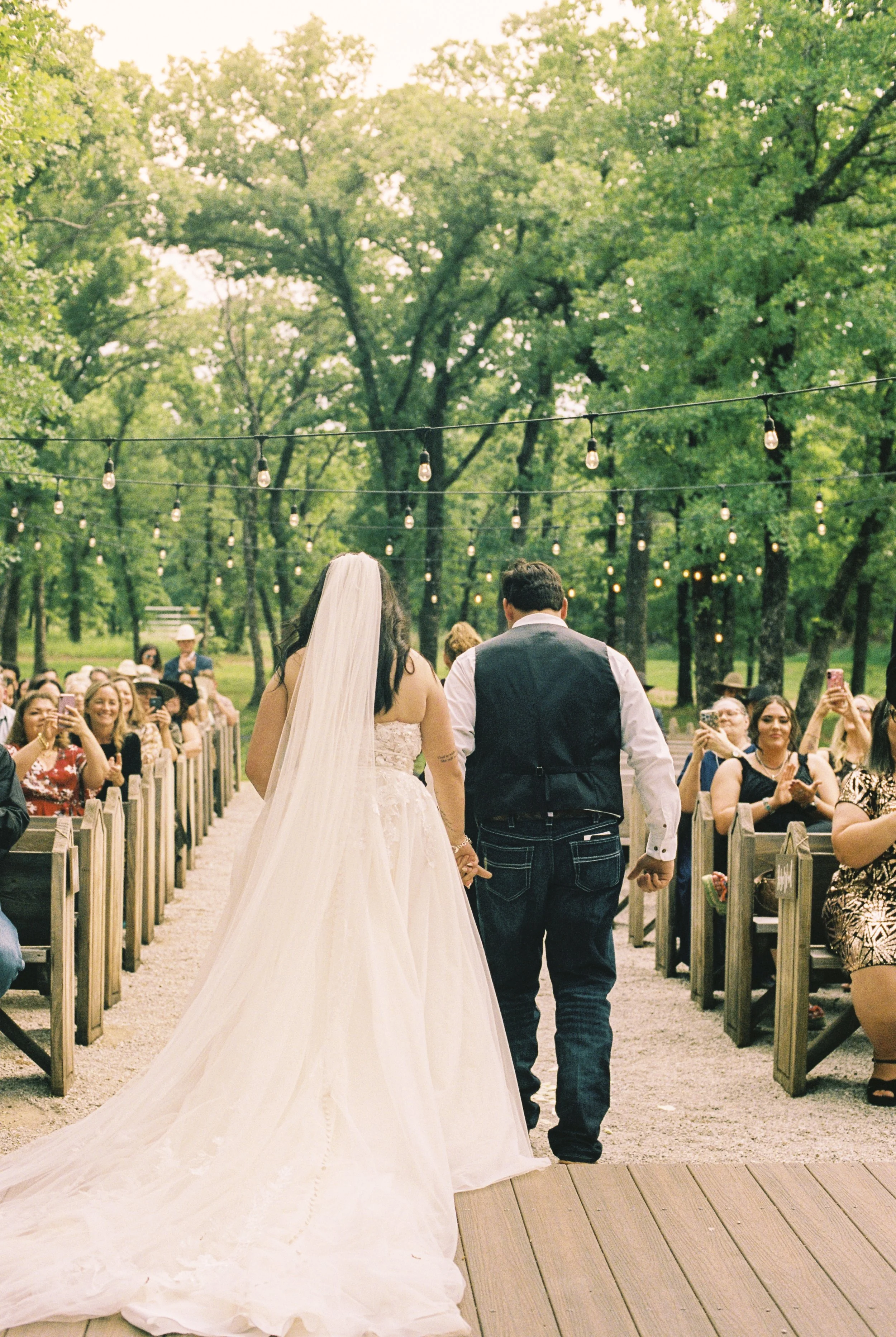 A bride and groom walking hand in hand down an outdoor aisle during a wedding ceremony with guests seated on both sides. The bride is wearing a white gown and veil, while the groom is dressed in a vest and shirt. String lights are hanging overhead am