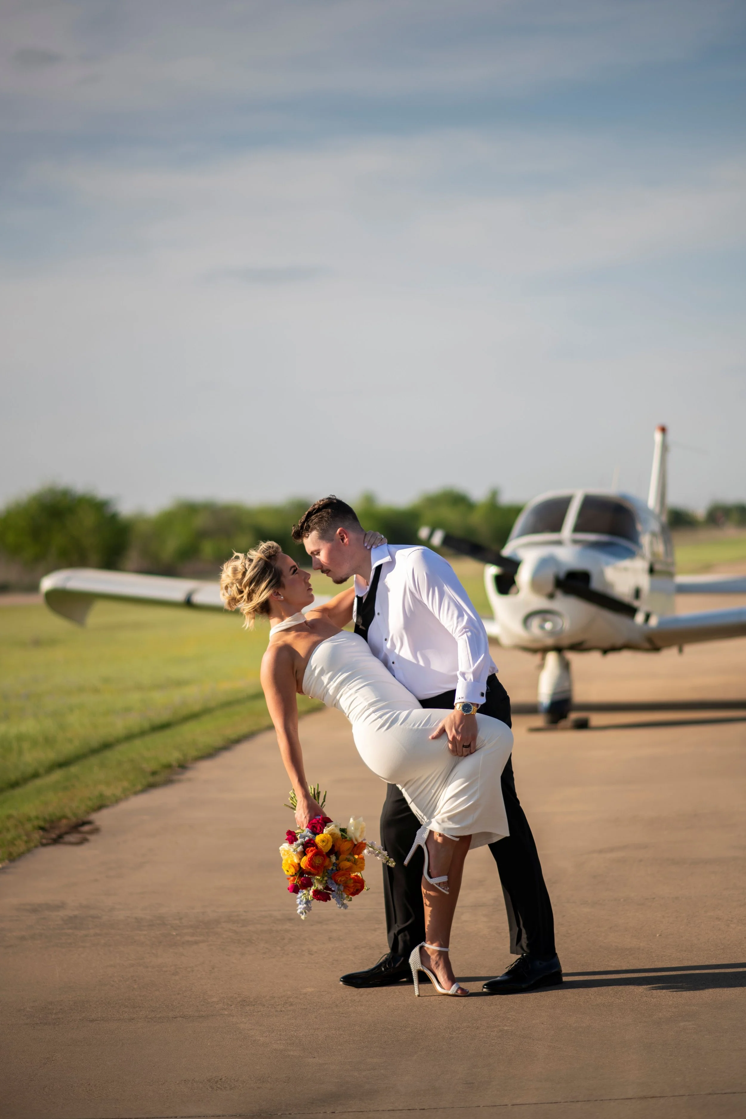 A man in a white shirt and black tuxedo pants is carrying a woman in a white wedding dress, who is holding a bouquet of colorful flowers. They are on an airport runway with a small private jet in the background.