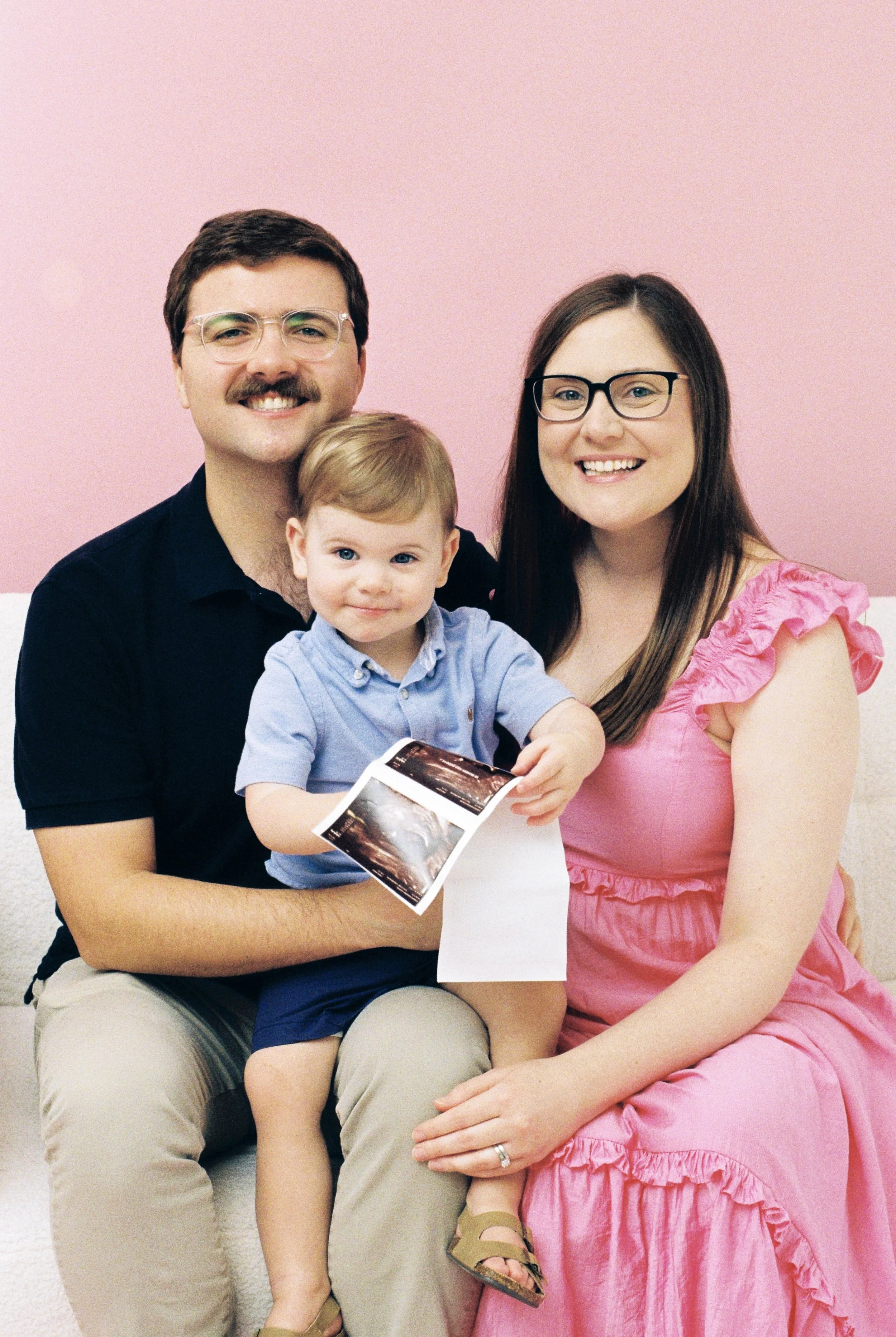 A family of three sitting on a couch, holding ultrasound images, with a pink wall background.