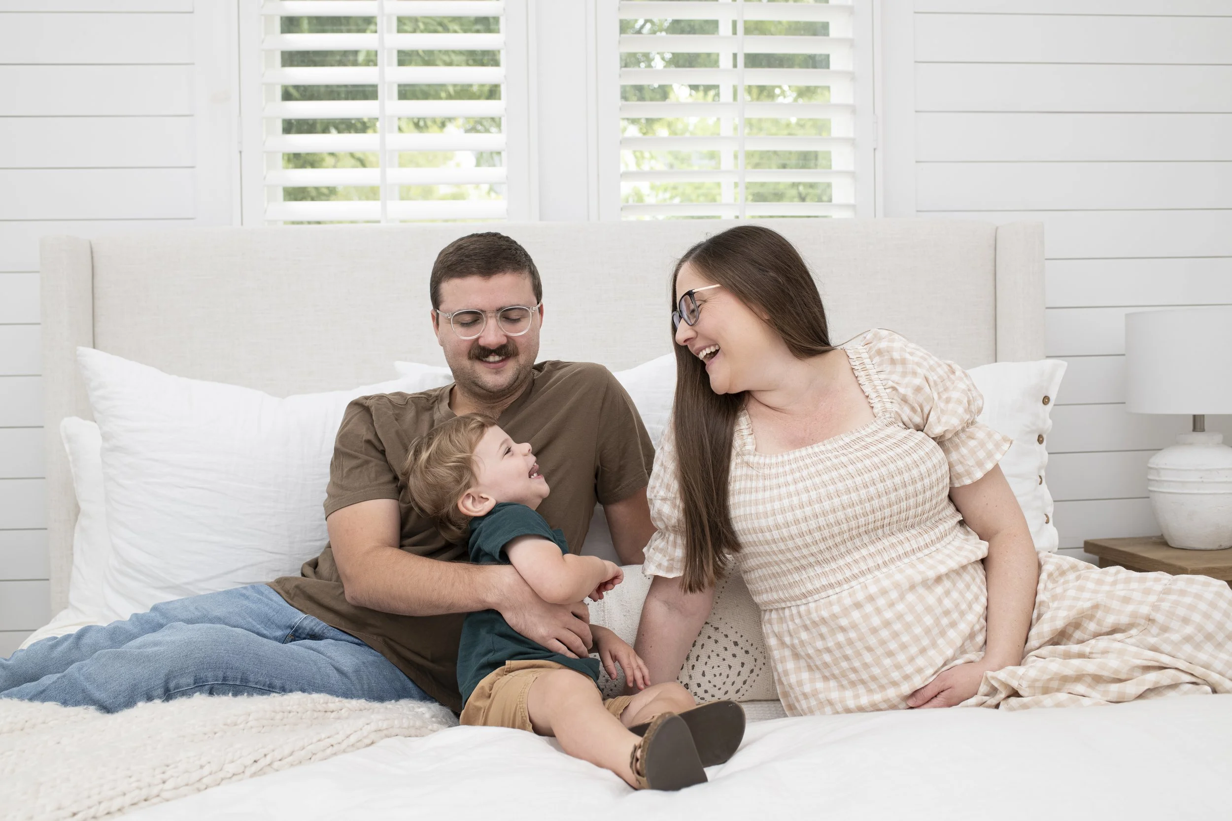 A family of three sitting on a bed and smiling at each other. The father is holding the young son, and the mother is leaning in, all appearing happy and engaged.