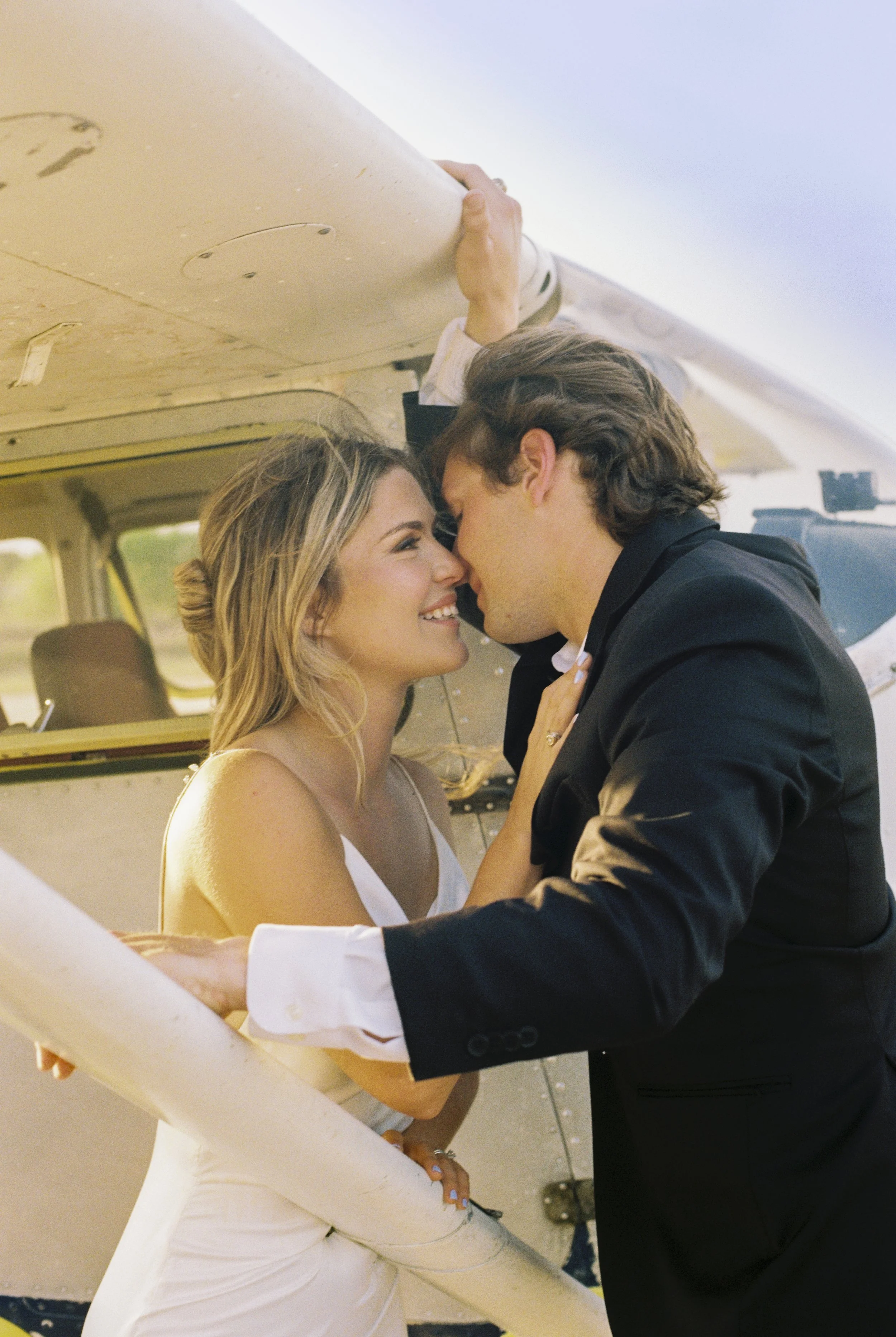A young couple, a woman in a white dress and a man in a black suit, share an intimate moment with their foreheads and noses touching, smiling at each other, inside a vehicle with light-colored walls and small windows.