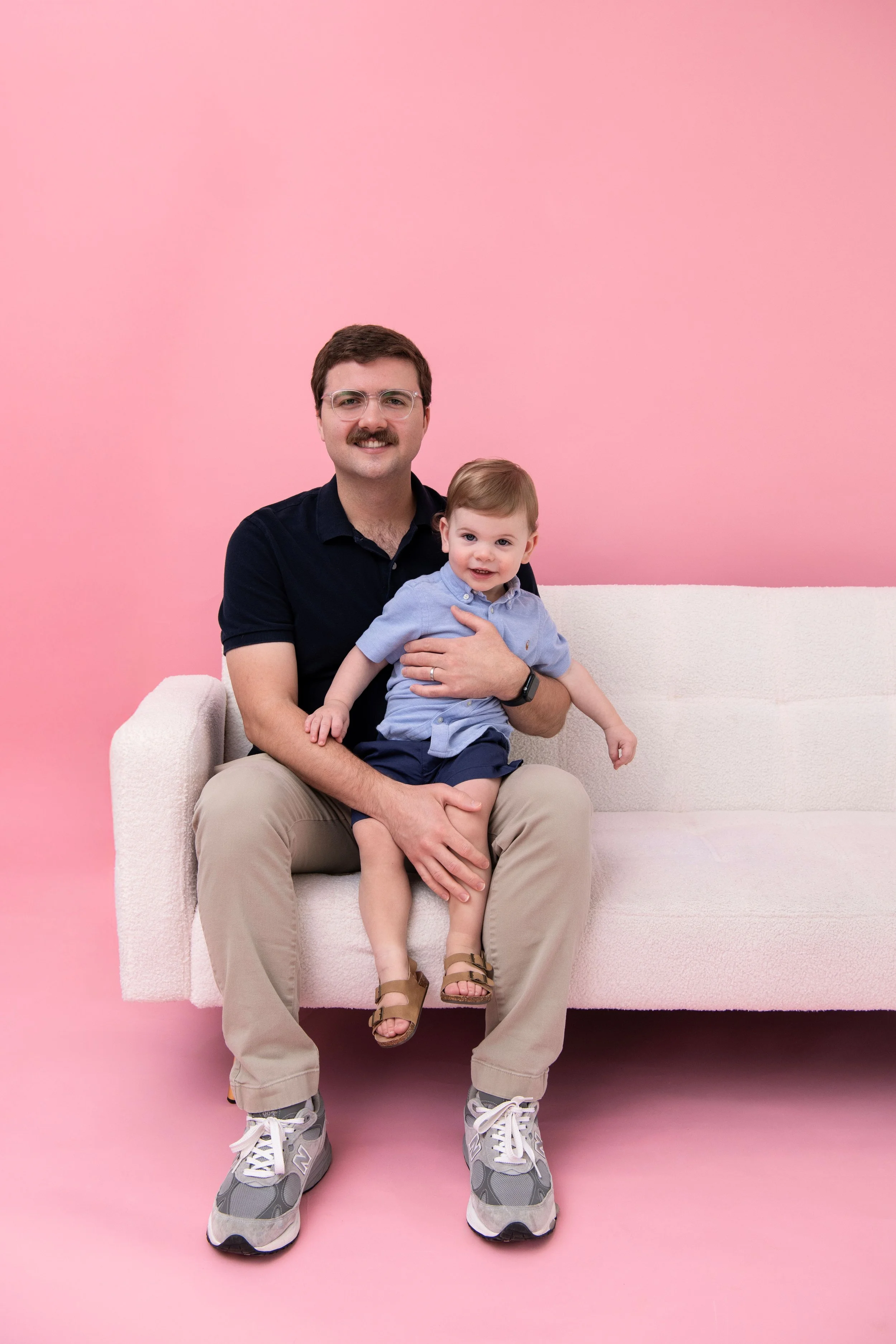 A man with glasses, a mustache, wearing a dark shirt and beige pants, is sitting on a white sofa holding a young boy in a blue shirt and shorts. They are smiling and sitting against a pink background.