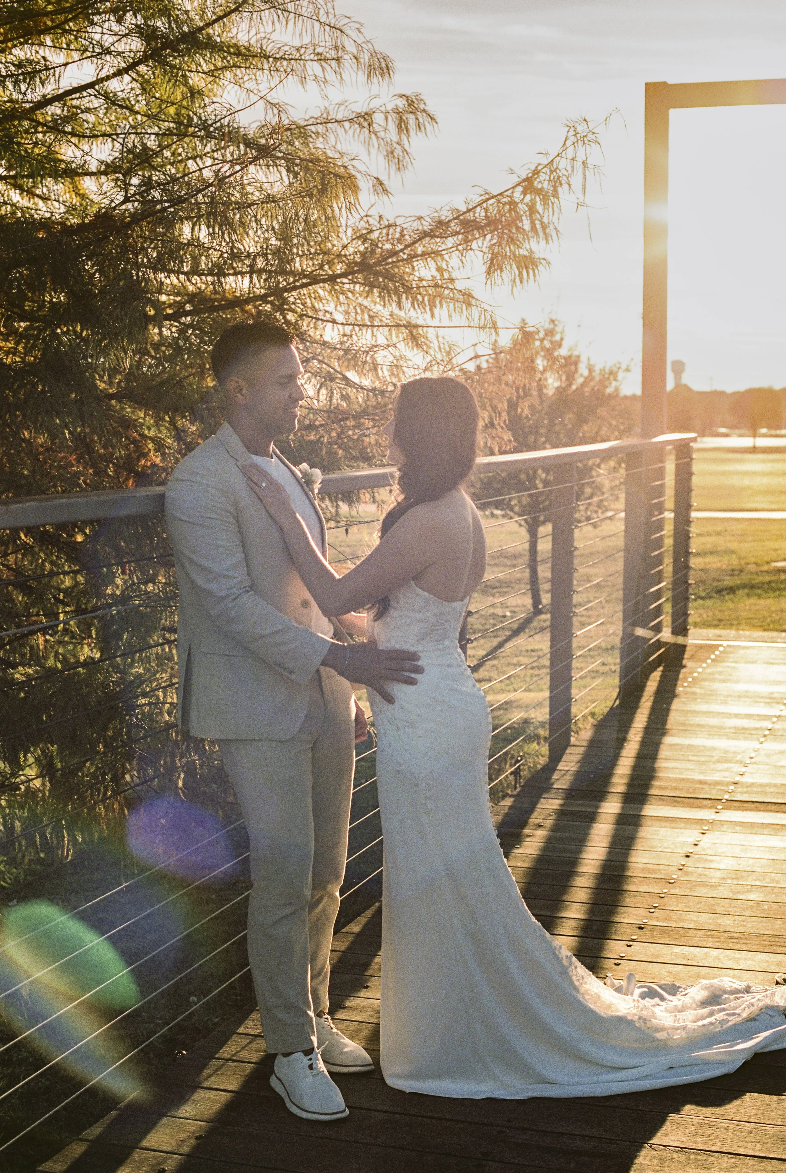 A newlywed couple sharing a romantic moment outdoors on a wooden bridge during sunset, with trees and a bright sky in the background.