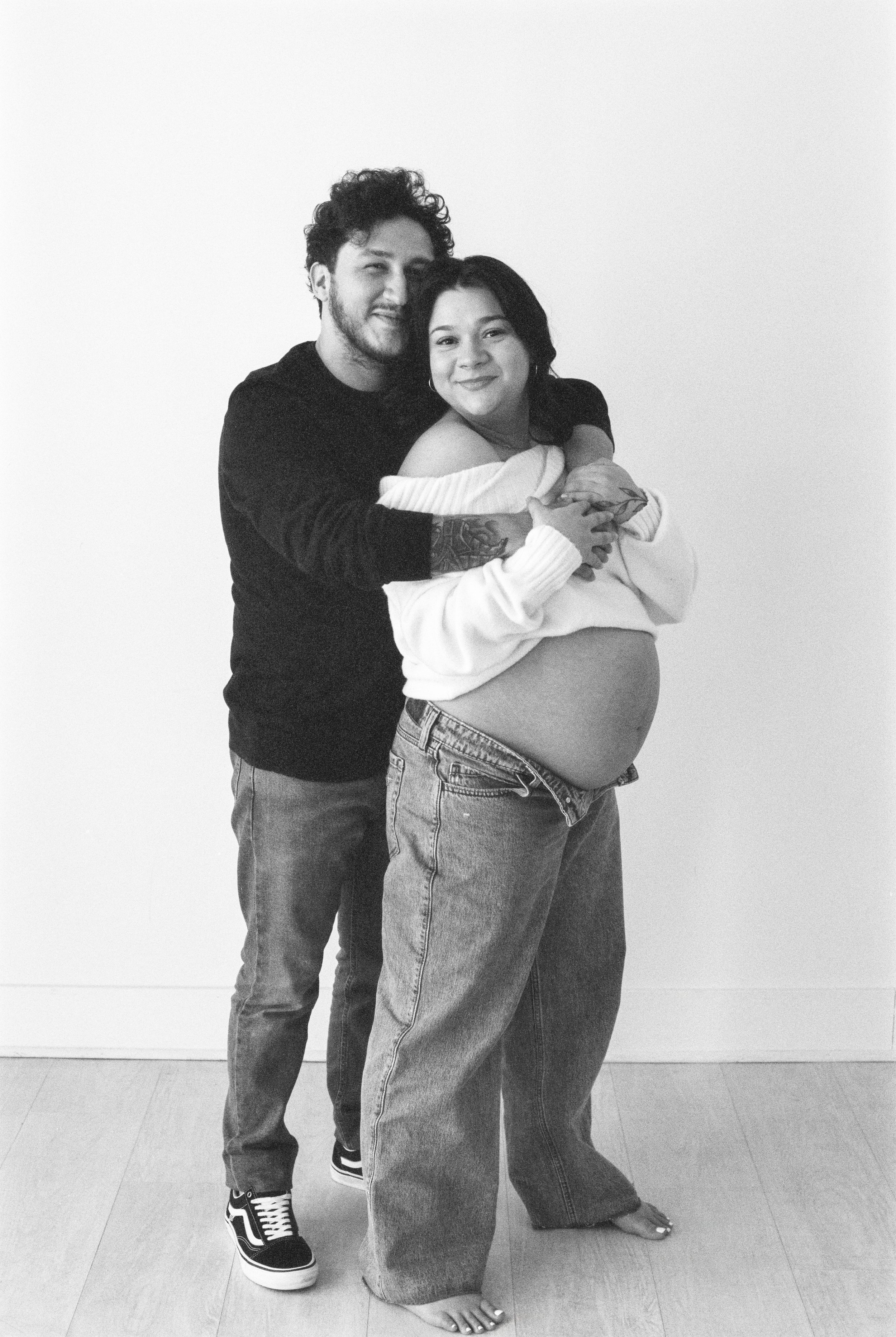 A man and a pregnant woman are standing together indoors against a plain wall, smiling and embracing each other. The woman is cradling her baby bump with one hand, and the man is holding her with both arms. The woman is wearing an off-the-shoulder sw