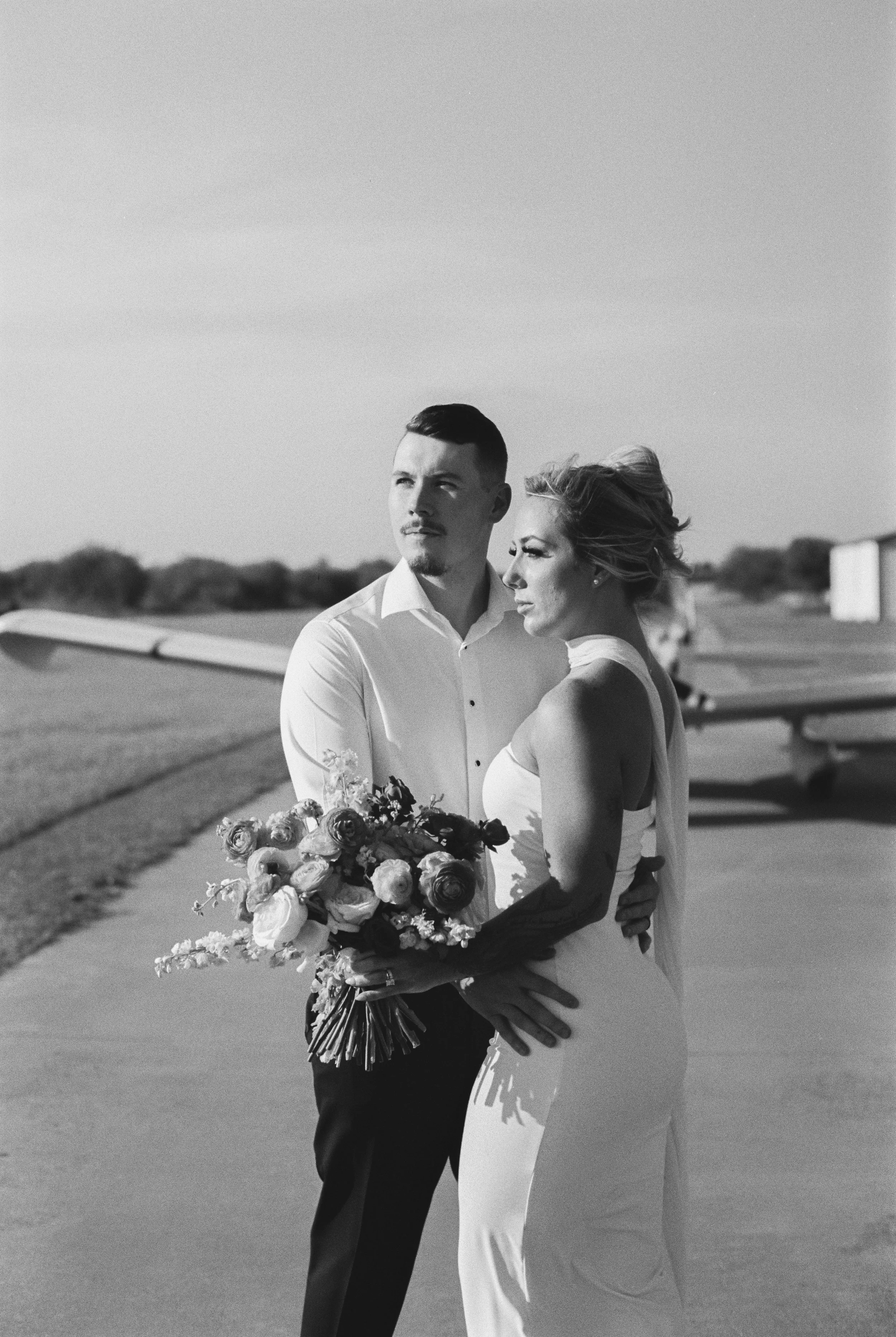 A black and white photo of a man and woman in wedding attire standing outdoors. The man is wearing a white shirt and dark pants, holding a bouquet of flowers. The woman is in a white gown, with her hair styled up. They are looking into the distance, 