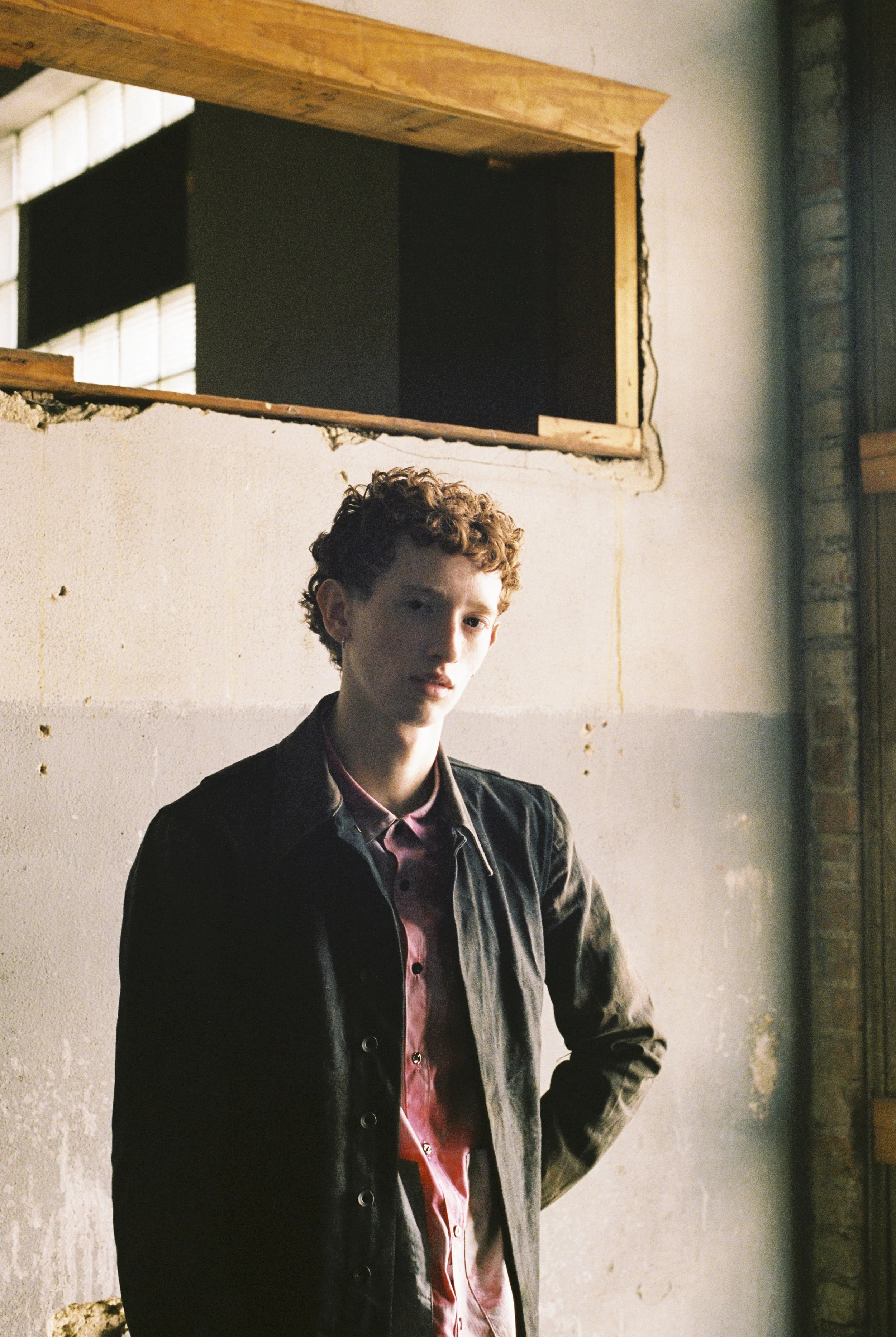 Young person with short curly hair wearing a black jacket and a pink shirt standing indoors near a wall with a large window in the background.