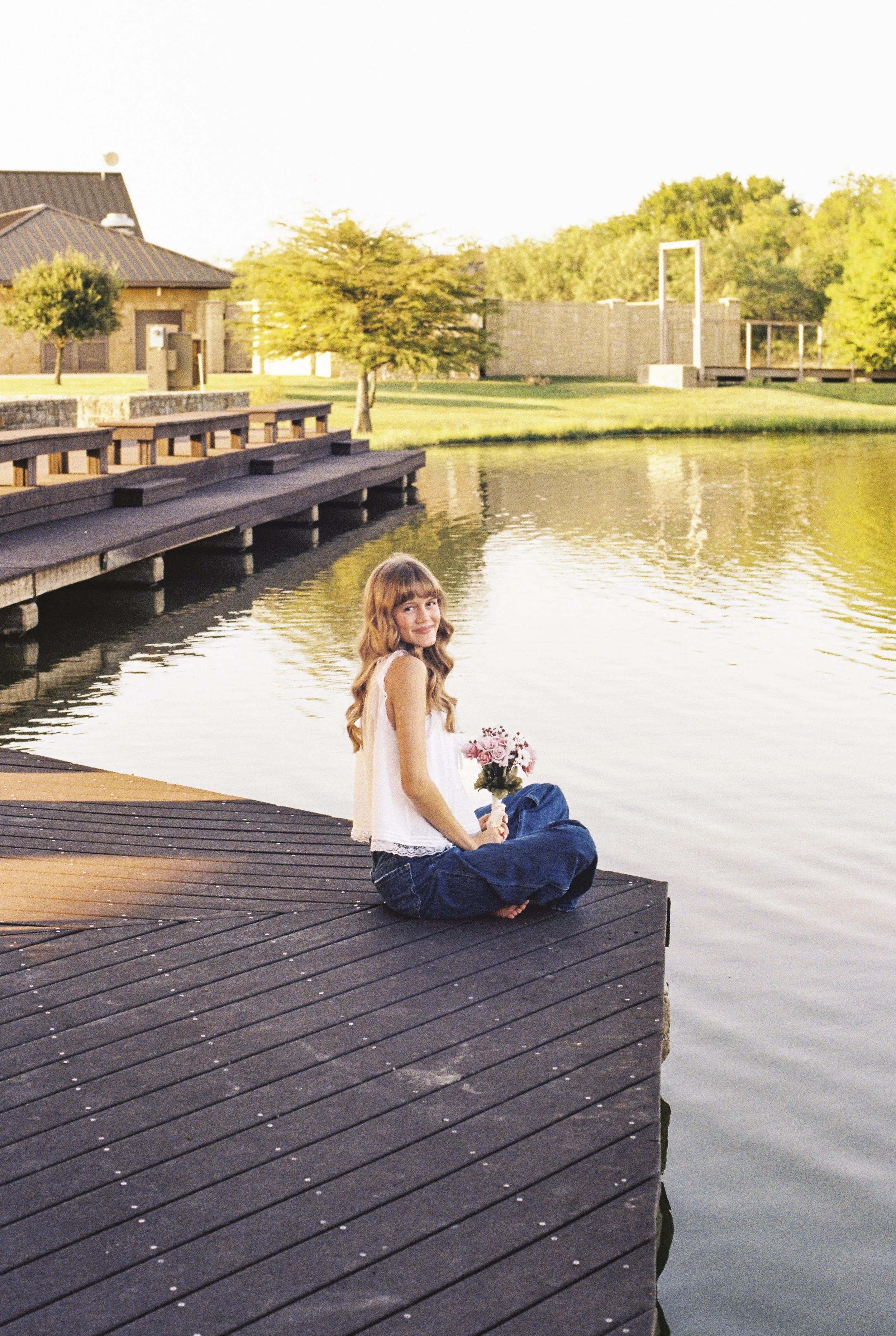 A young woman with long, wavy hair sitting on a wooden dock by a lake, holding a bouquet of flowers, with trees and houses in the background during sunset.